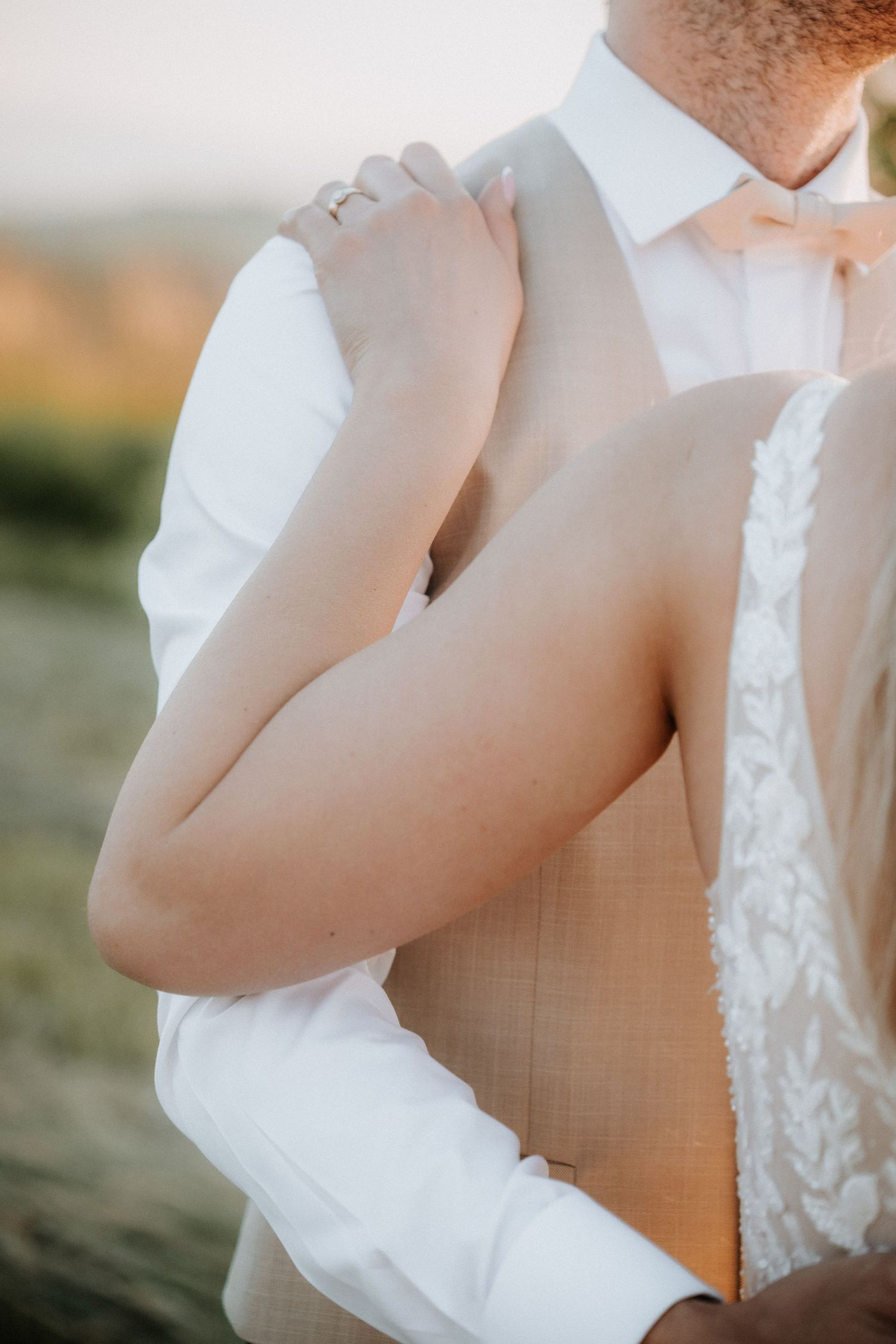 Bride and groom embracing outdoors, bride wearing lace wedding dress with groom in white shirt and beige vest at sunset.