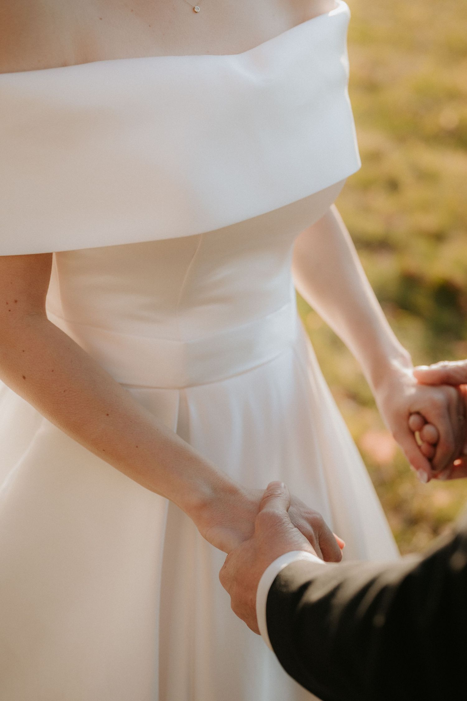 Bride in white off-shoulder gown holding hands with groom in black suit outdoors on wedding day.