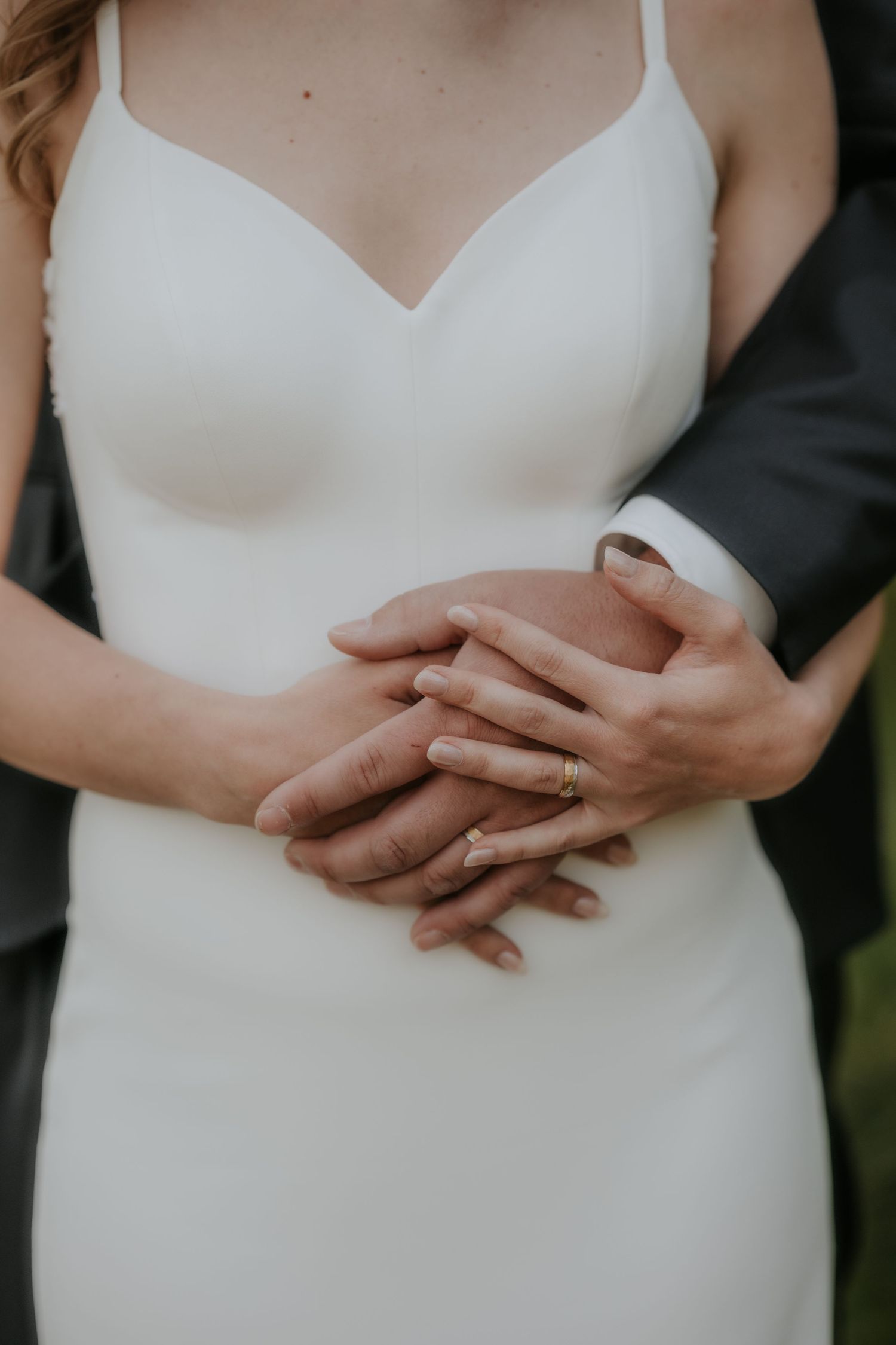 Bride and groom holding hands together, showcasing a gold wedding ring, with bride in elegant white spaghetti strap gown.