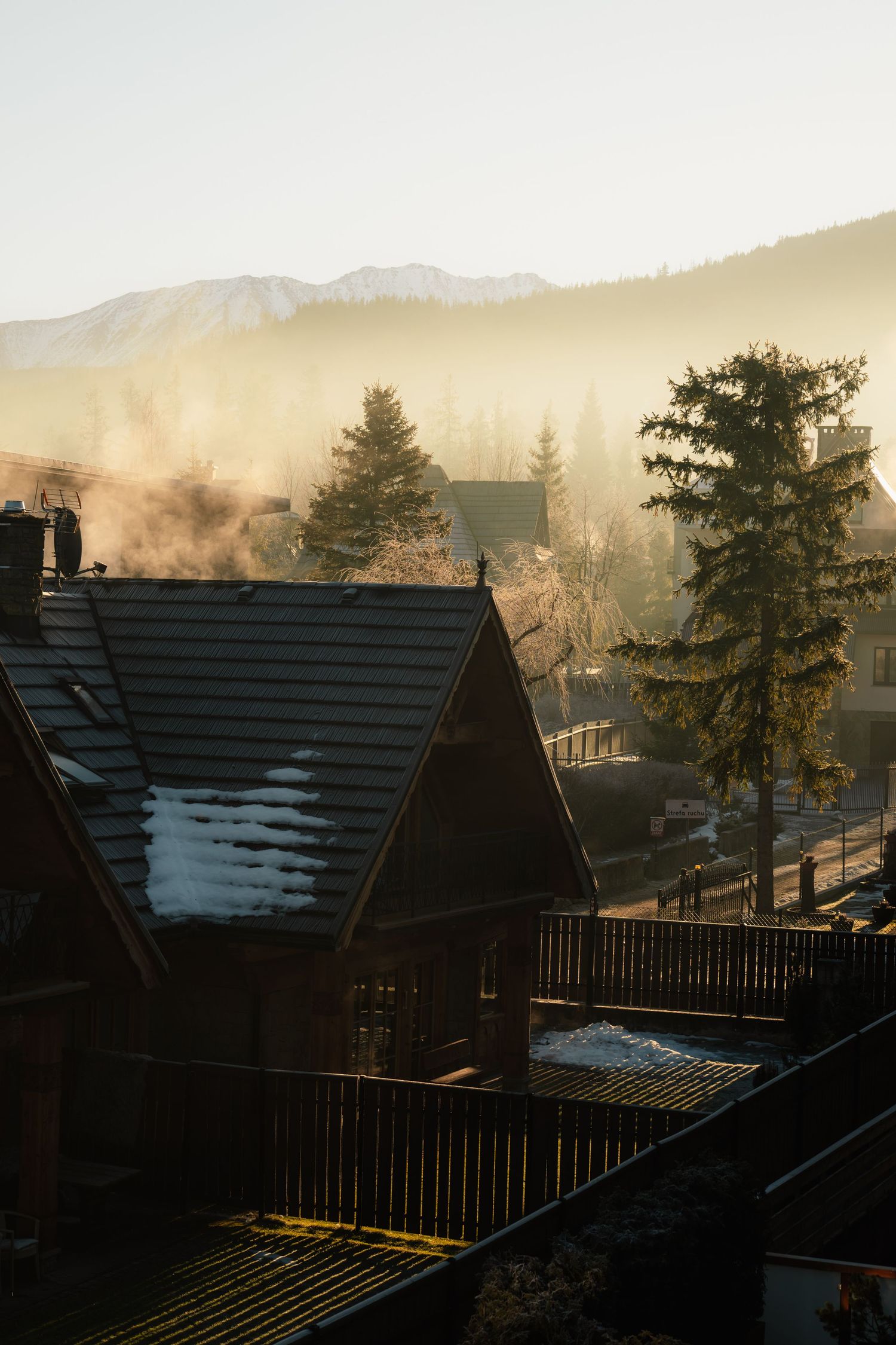 Wooden mountain chalet with snow-dusted roof emitting chimney smoke at golden sunrise with misty pine forest backdrop.