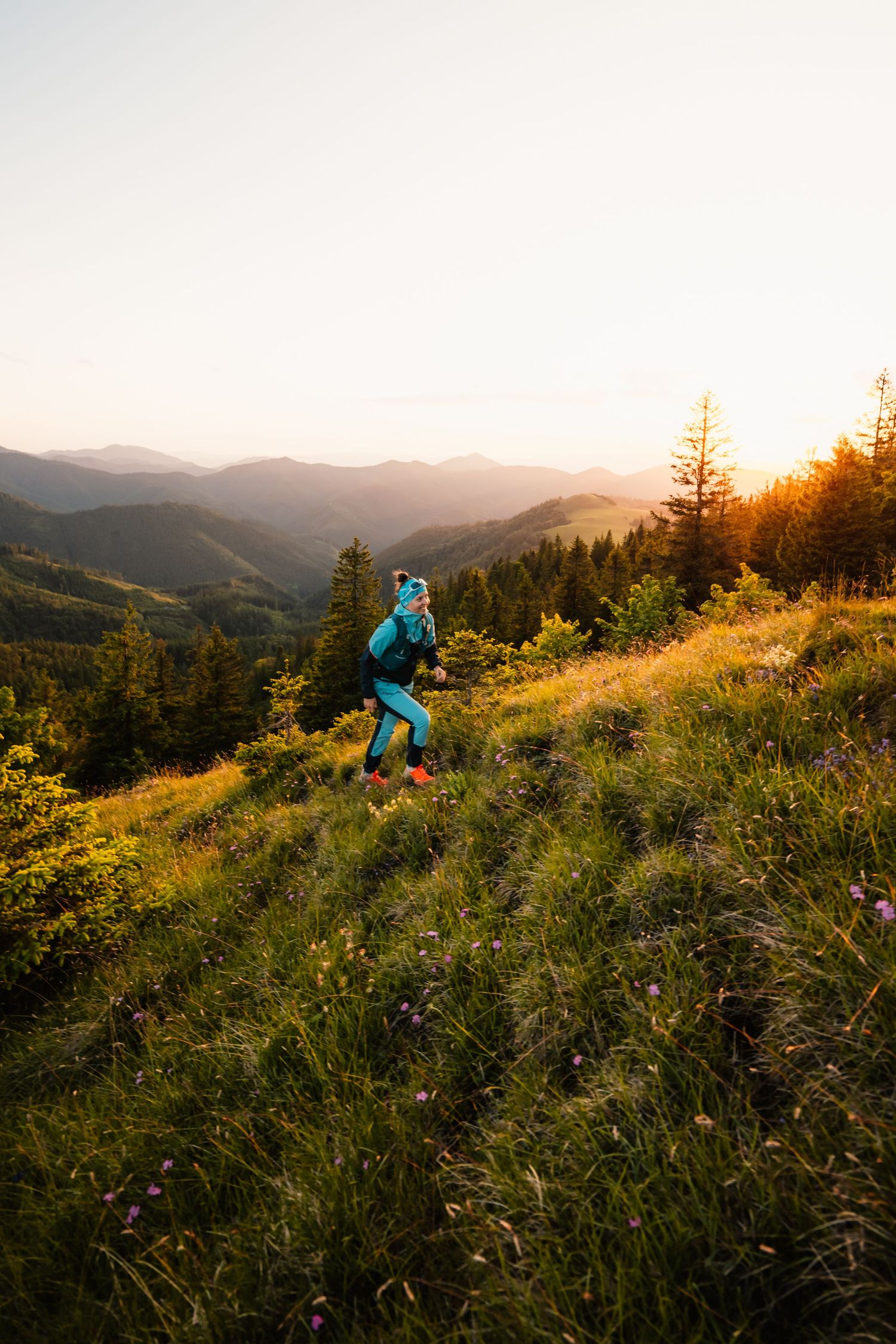 A trail runner in teal athletic gear runs through a wildflower mountain meadow at golden sunset.