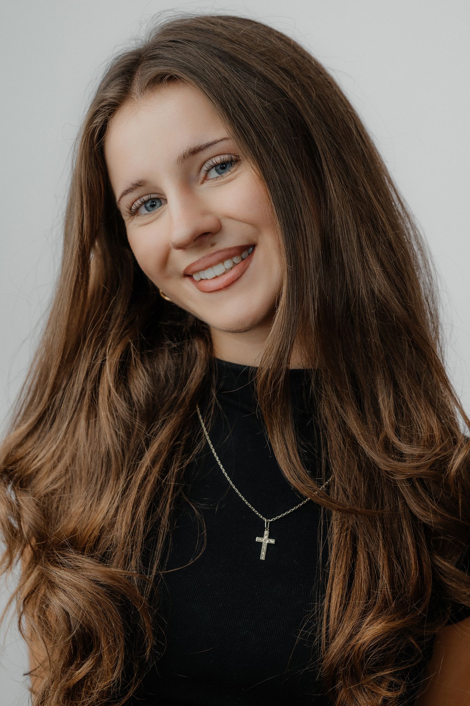 Young woman with long wavy brown hair wearing a cross necklace and black top, smiling with braces, gray background.