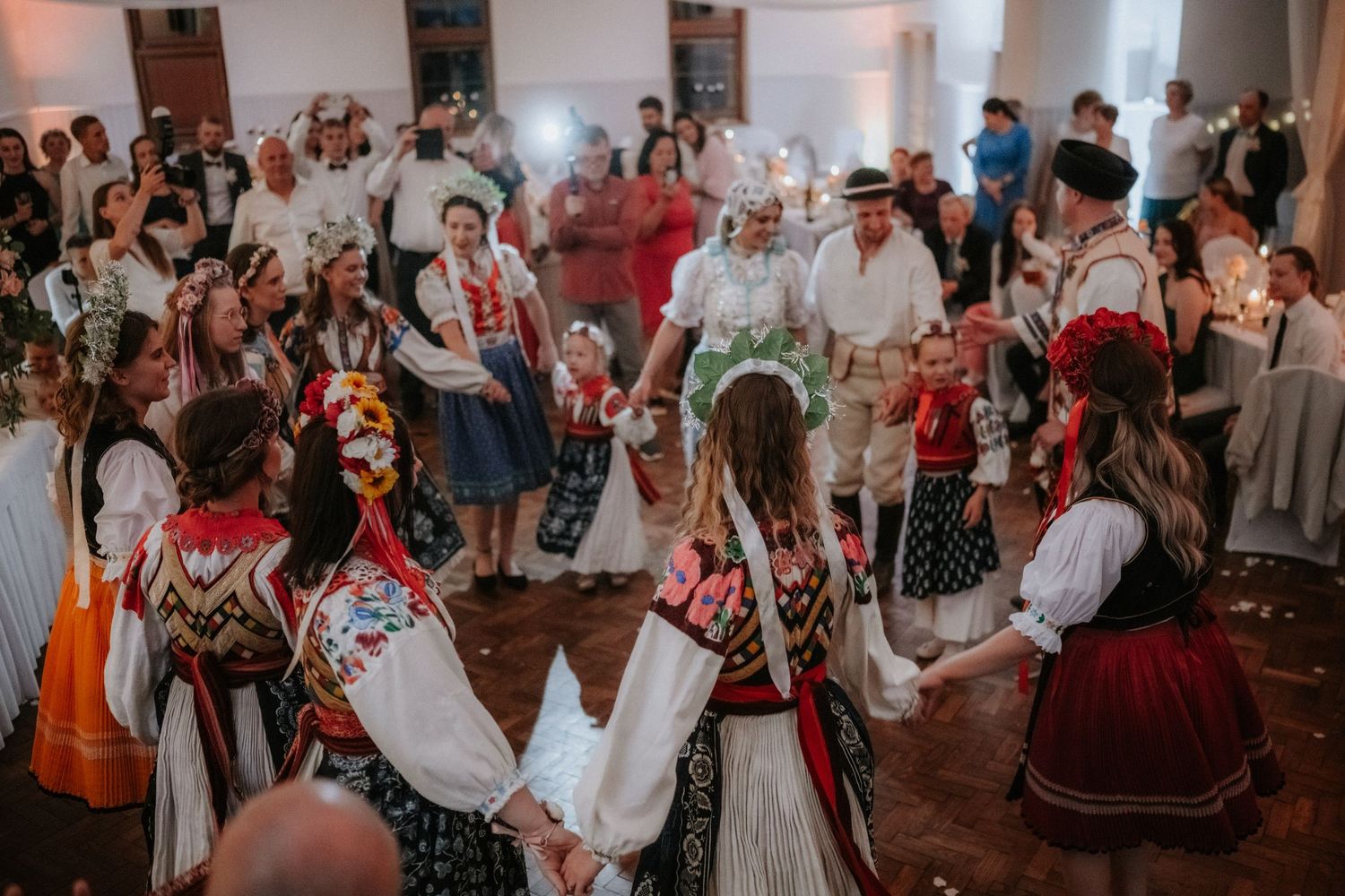 Traditional Polish folk dancers in colorful embroidered costumes performing a circle dance at a festive wedding celebration.