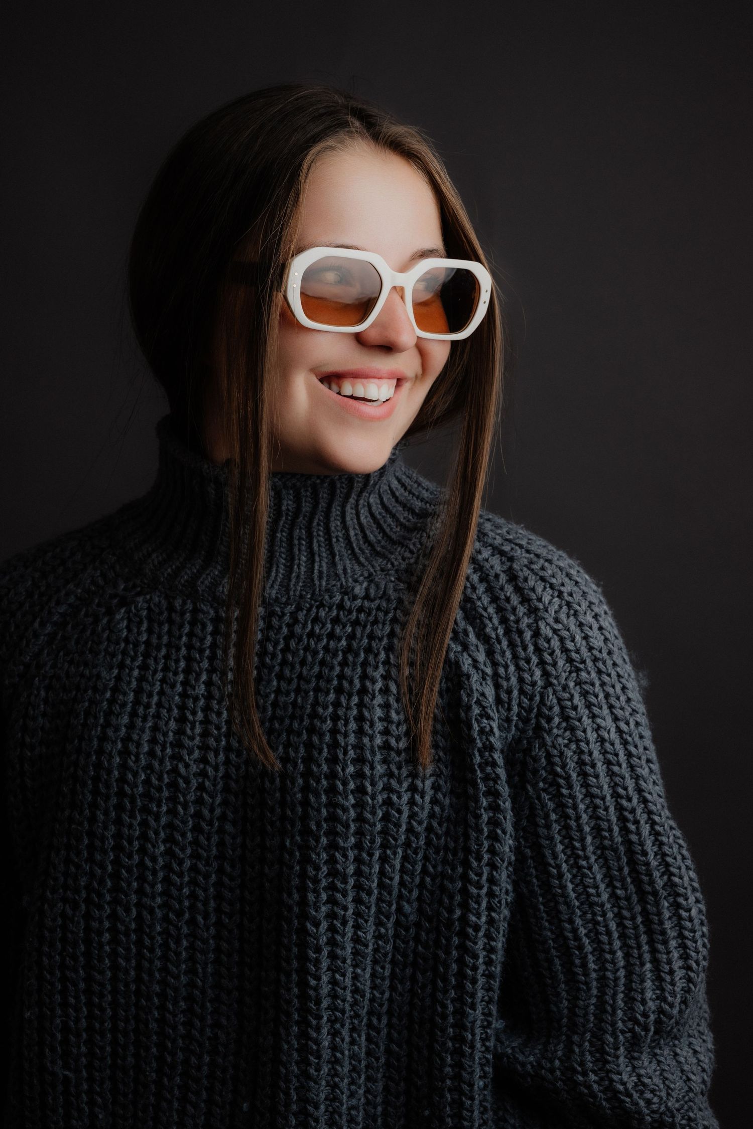 Smiling young woman wearing oversized white square sunglasses and a chunky grey turtleneck sweater on a dark background.