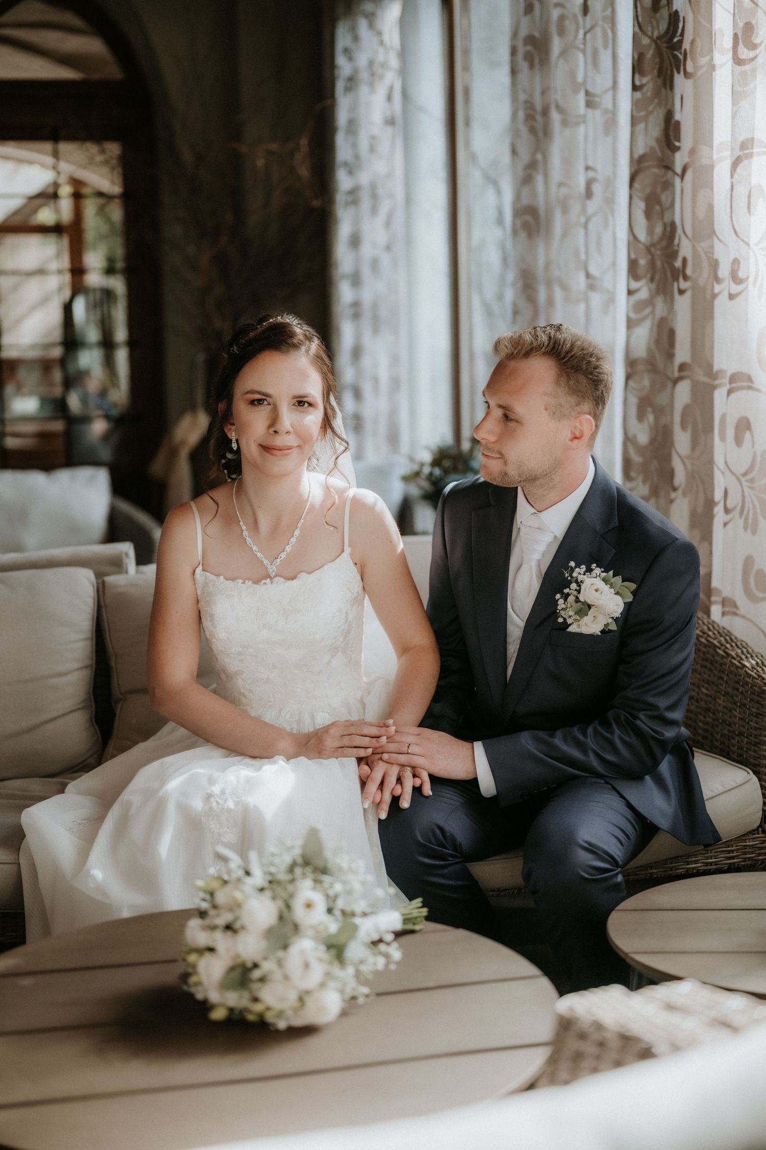 Bride and groom sitting together on a sofa holding hands with wedding bouquet in foreground.