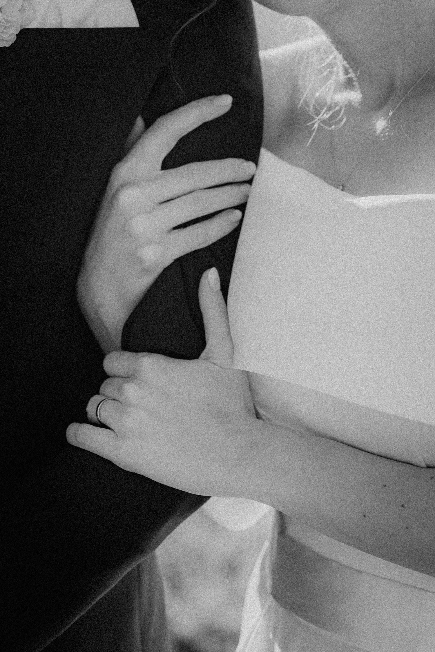 Black and white photo of a couple embracing, showing wedding rings on their hands, bride in white dress.