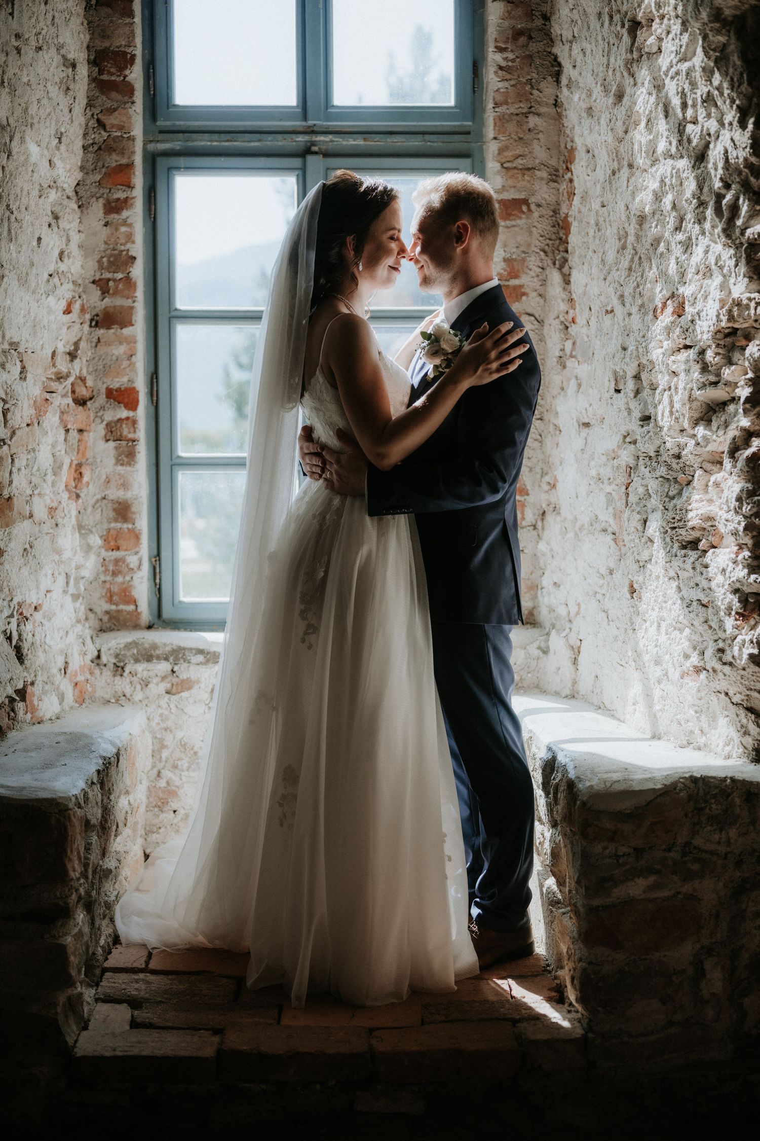 Bride and groom embracing near a rustic stone window in romantic wedding portrait.