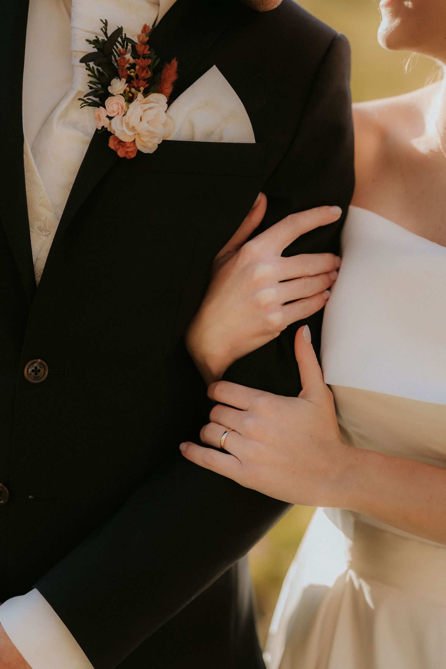 Bride in white dress embracing groom in black tuxedo with floral boutonniere, wedding ring visible on her hand.