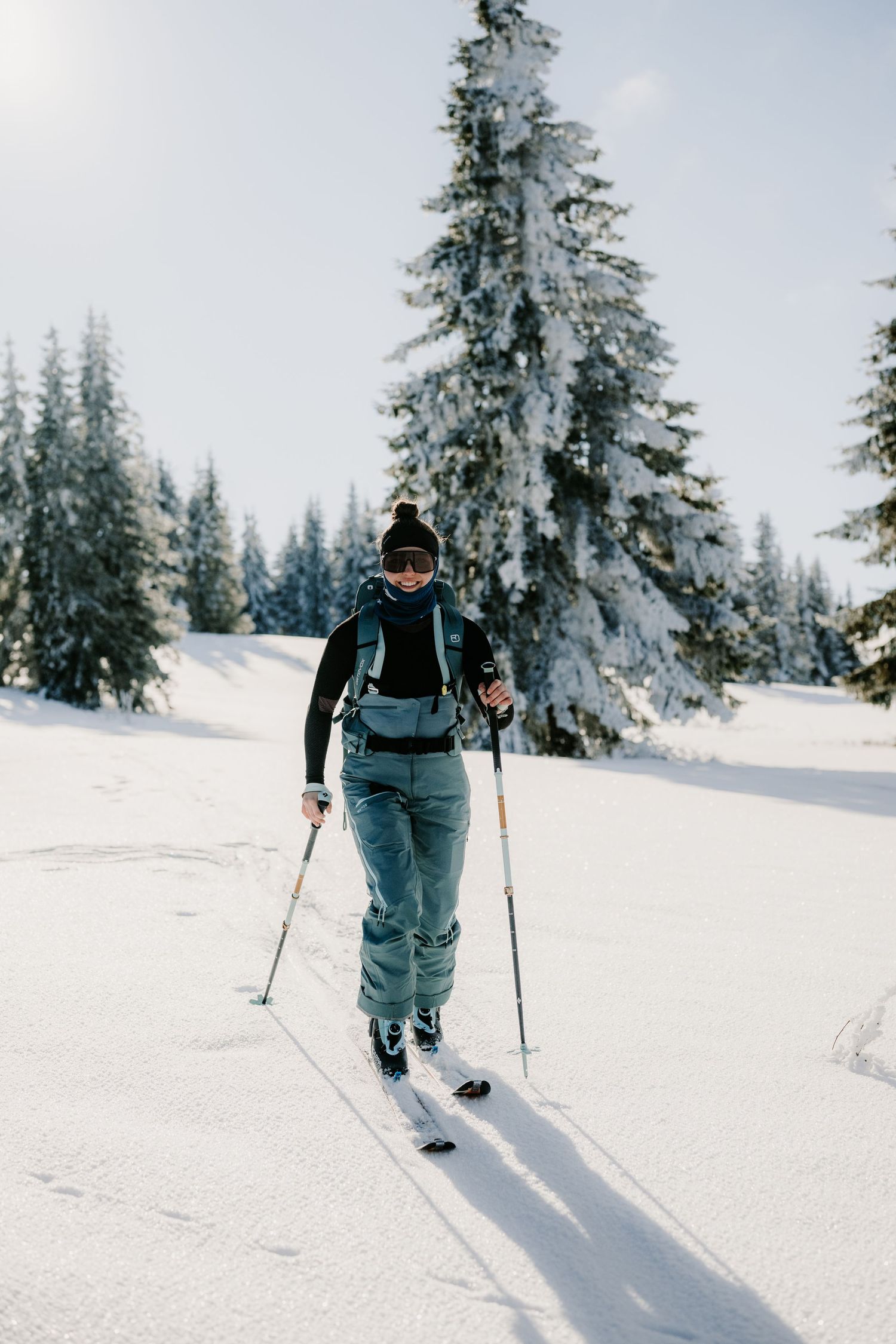 A solo skier glides across a pristine snow-covered landscape flanked by frost-covered pine trees.