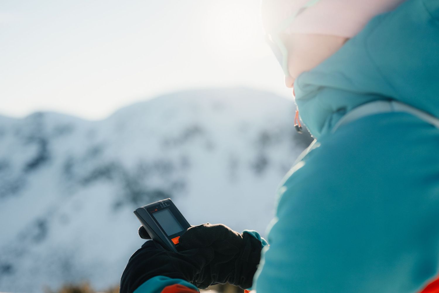 Close-up of skier in teal jacket using handheld device with sun-lit snowy mountain in background.