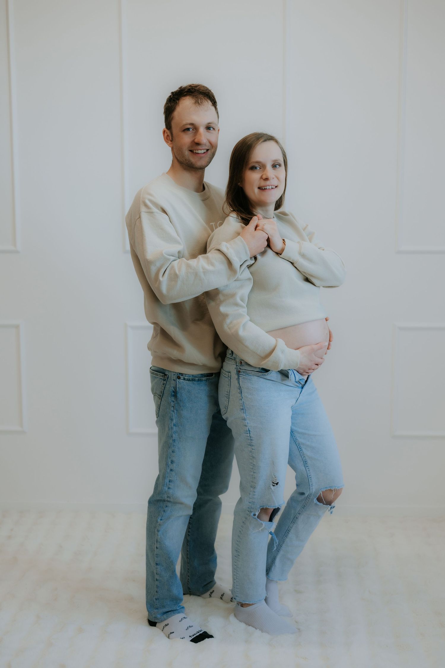 Couple in beige sweatshirts embrace as man holds pregnant womans baby bump from behind.