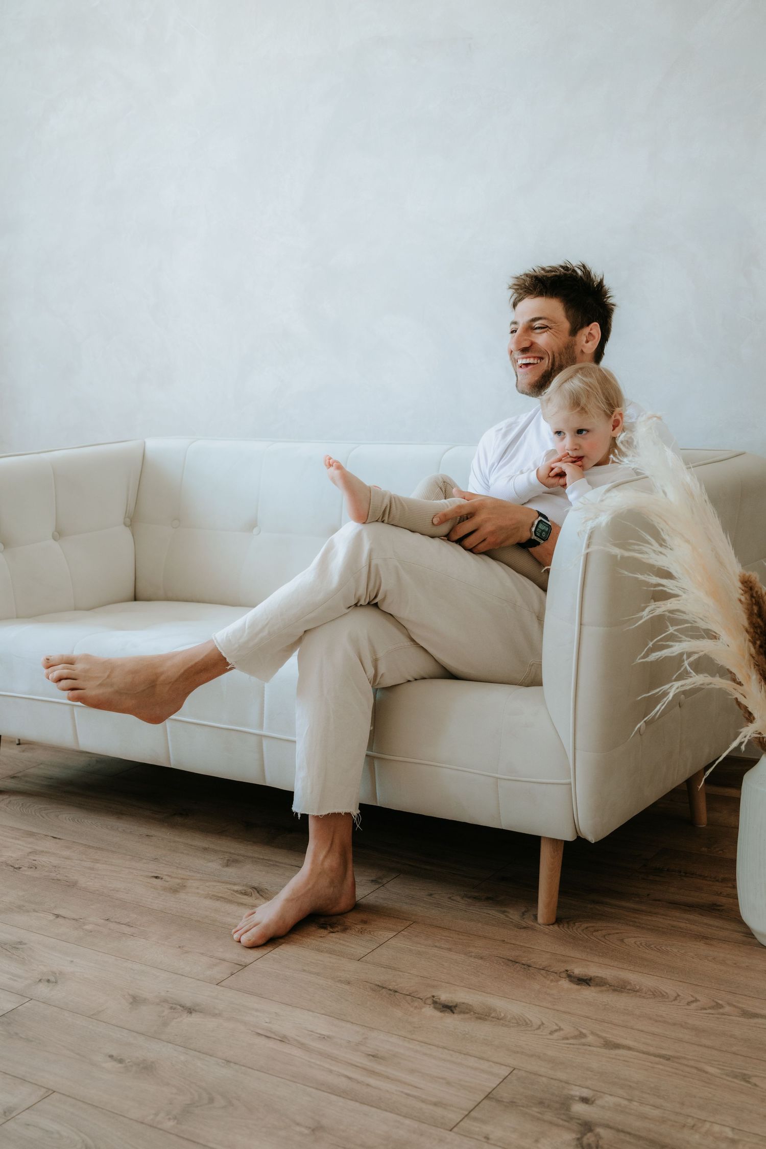 Happy father sitting on beige sofa laughing with baby girl in bright modern living room.
