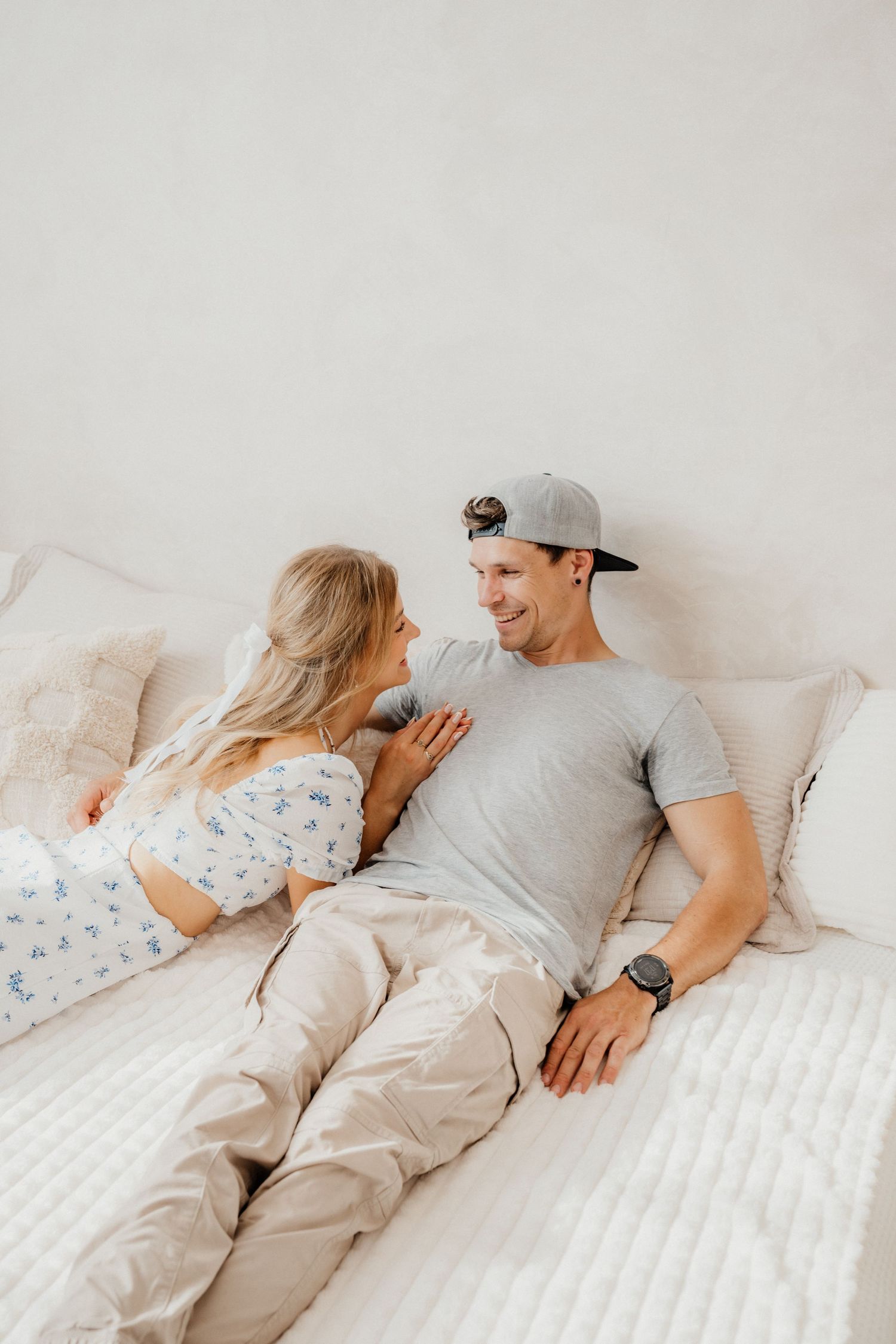A smiling couple reclines on a white bed, gazing at each other in a soft neutral-toned bedroom environment.