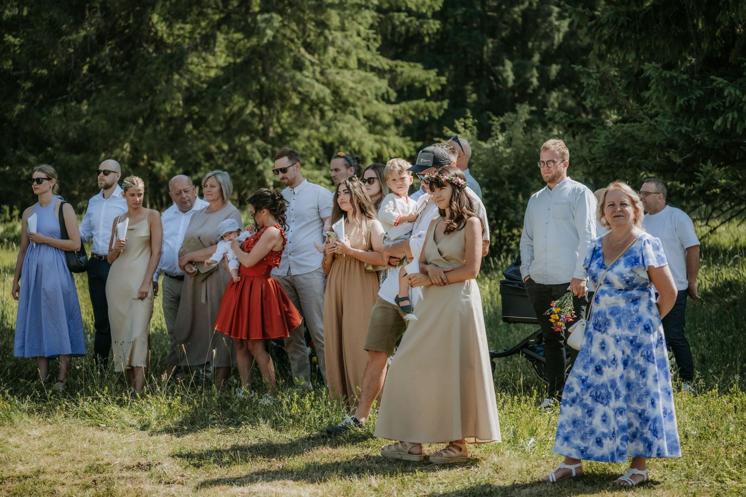 Wedding guests gathered outdoors on green grass, dressed in elegant attire, celebrating a summer ceremony surrounded by trees.