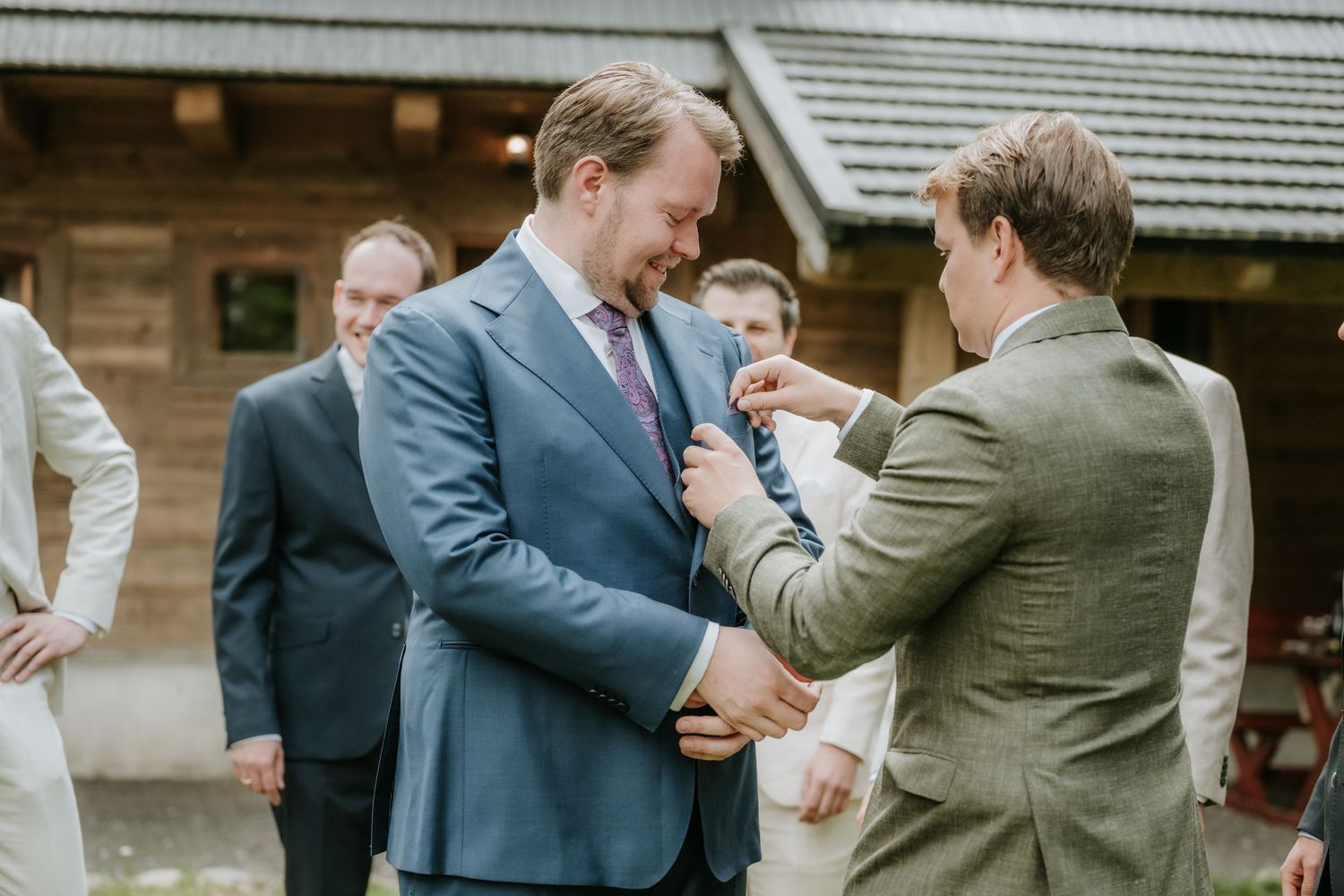 Groom in blue suit getting boutonniere pinned by groomsman outdoors at rustic wedding venue.