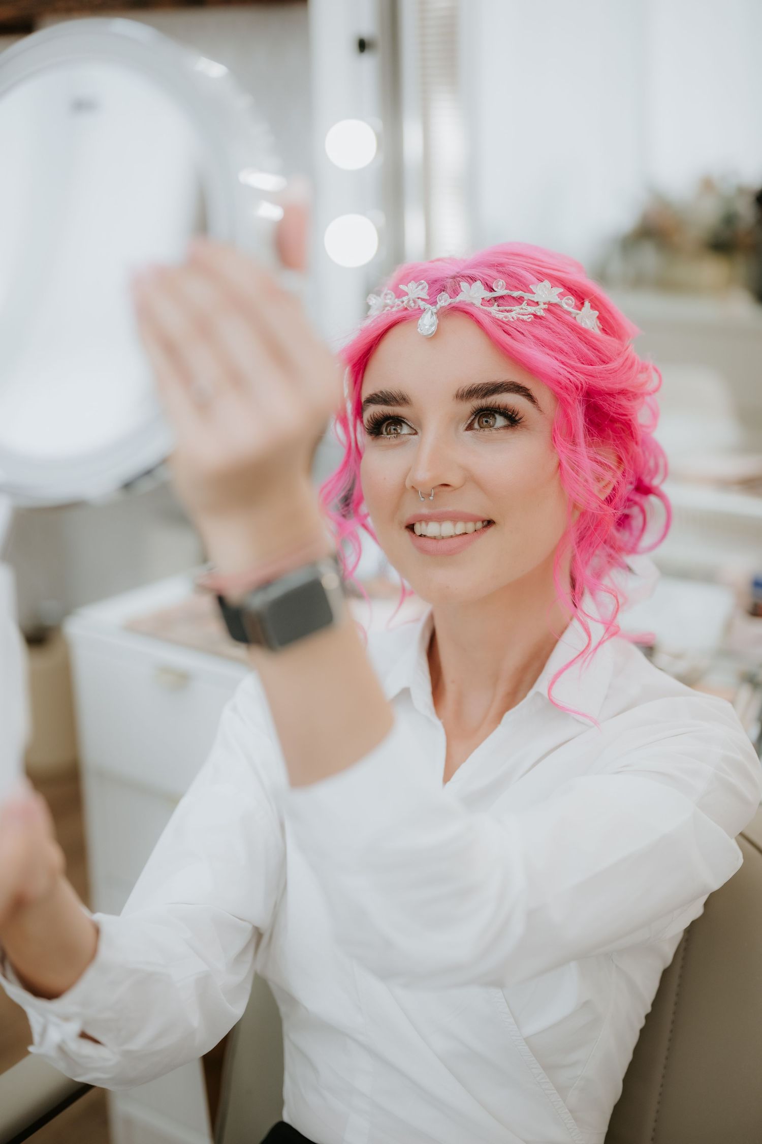 Woman with pink curly hair and tiara holding a mirror, wearing a white shirt, smiling in a bright salon.