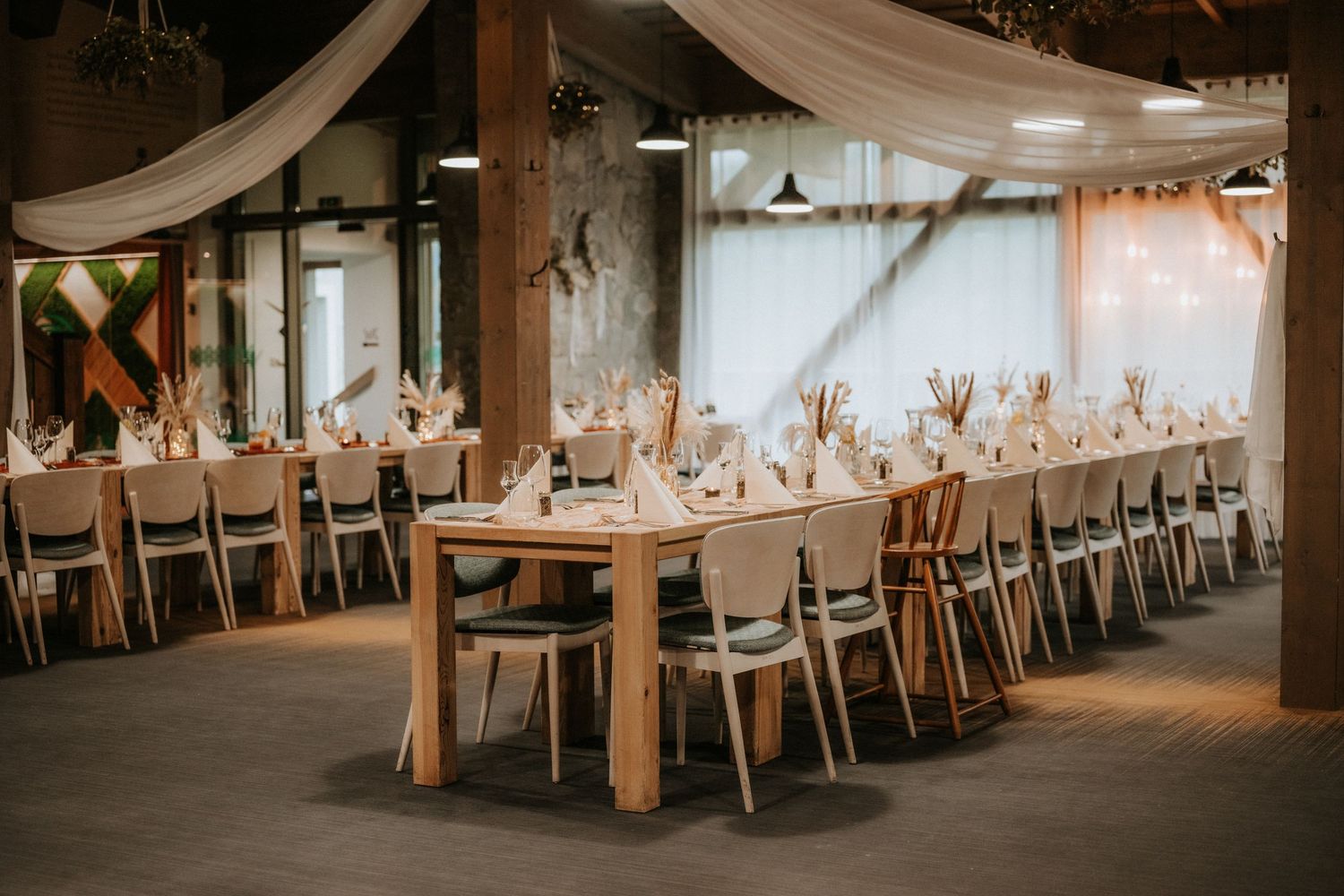 Elegant wedding reception hall with long wooden tables, white chairs, and draped fabric ceiling decor.