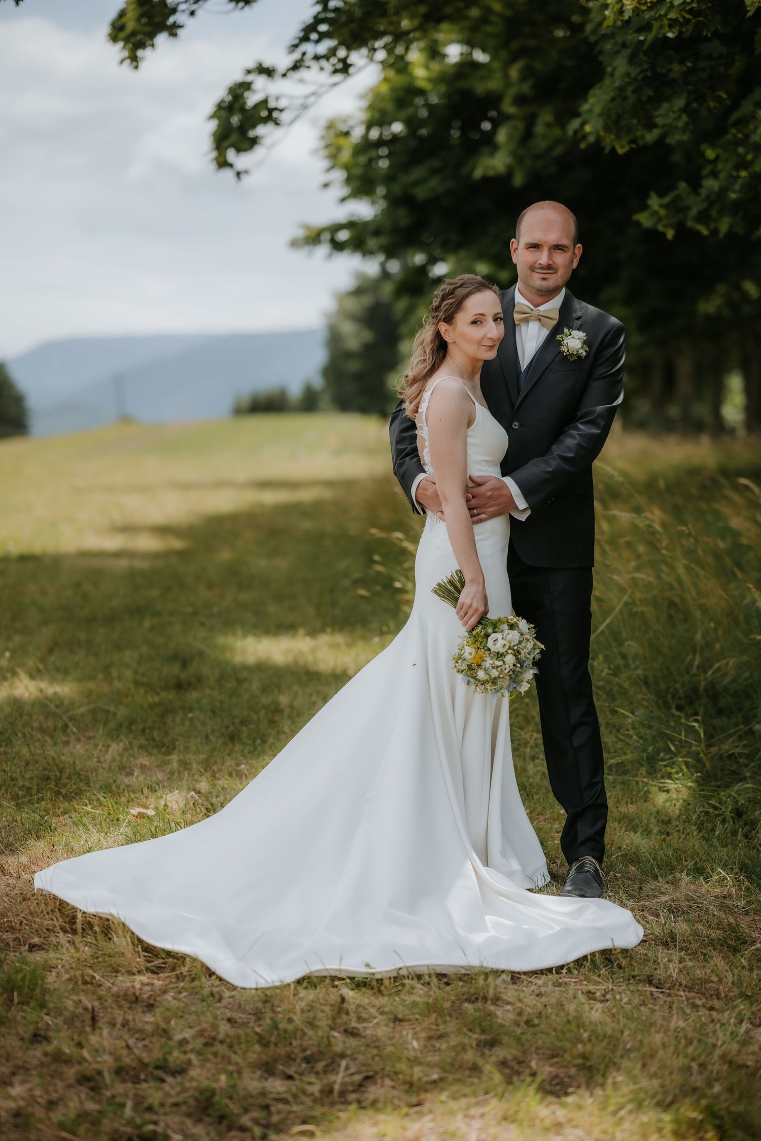 Bride in white gown holding bouquet stands with groom in dark suit on grassy path surrounded by trees.