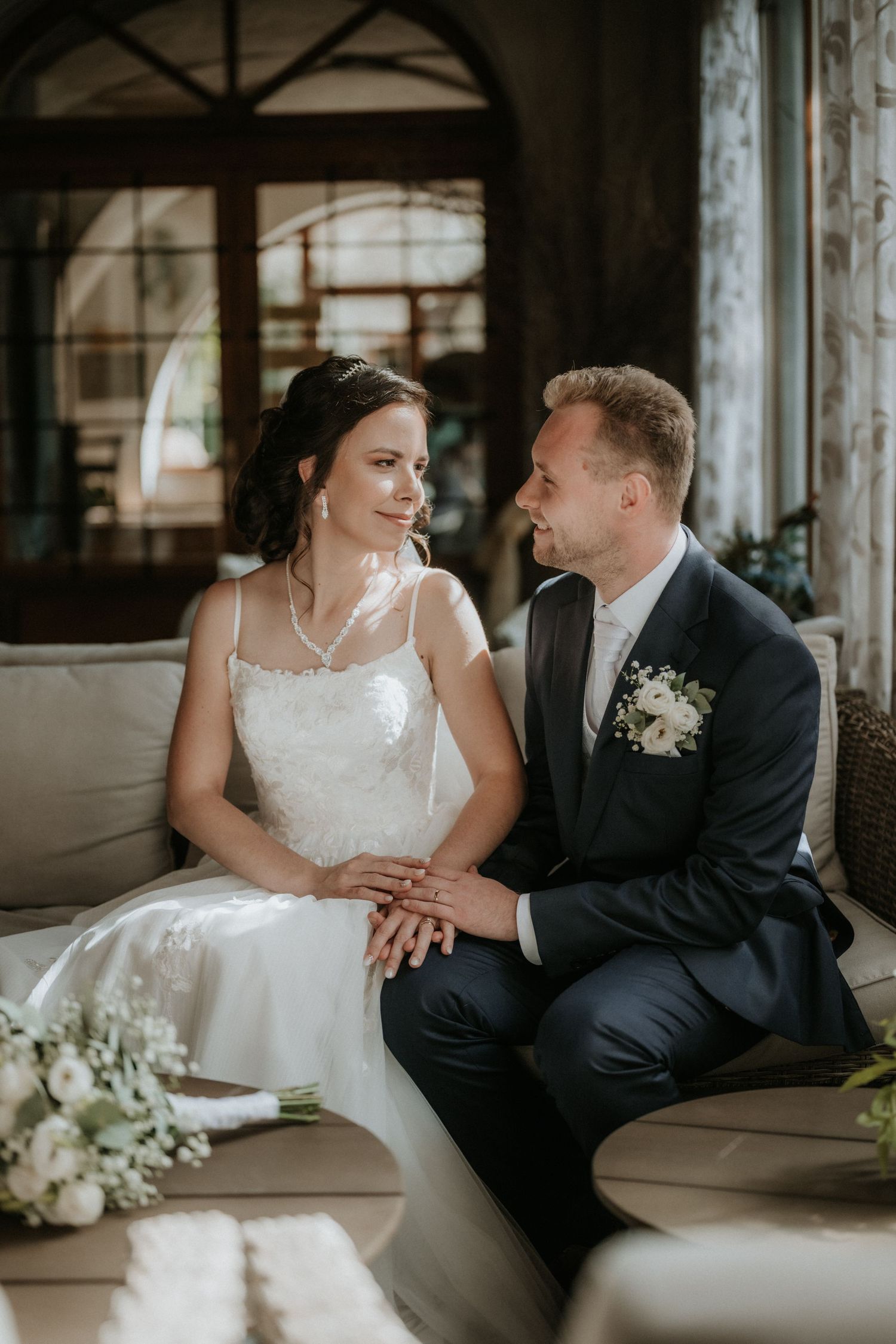 Bride and groom sitting together on a couch, holding hands, dressed in wedding attire in an elegant indoor setting.