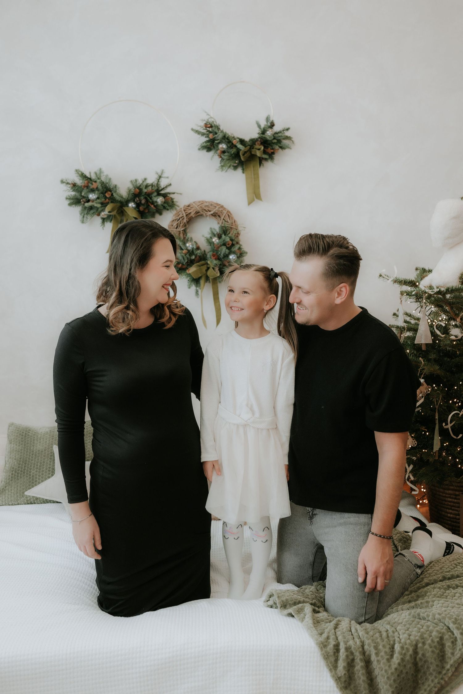 A happy family of three poses in front of Christmas wreaths, mom in black dress, dad in black tee, daughter in white dress.