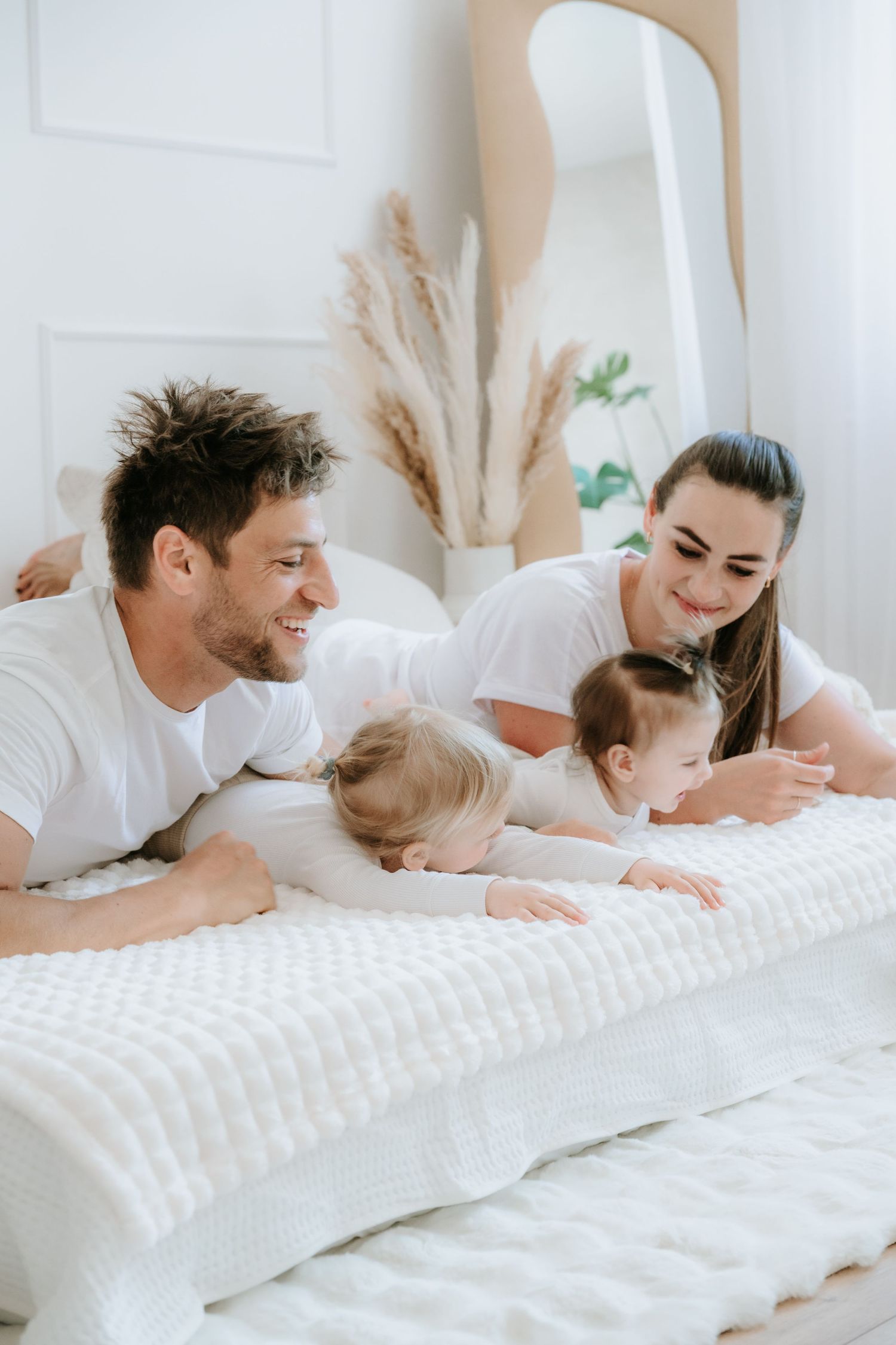 Smiling parents in white shirts lying on bed watching their two young children play.