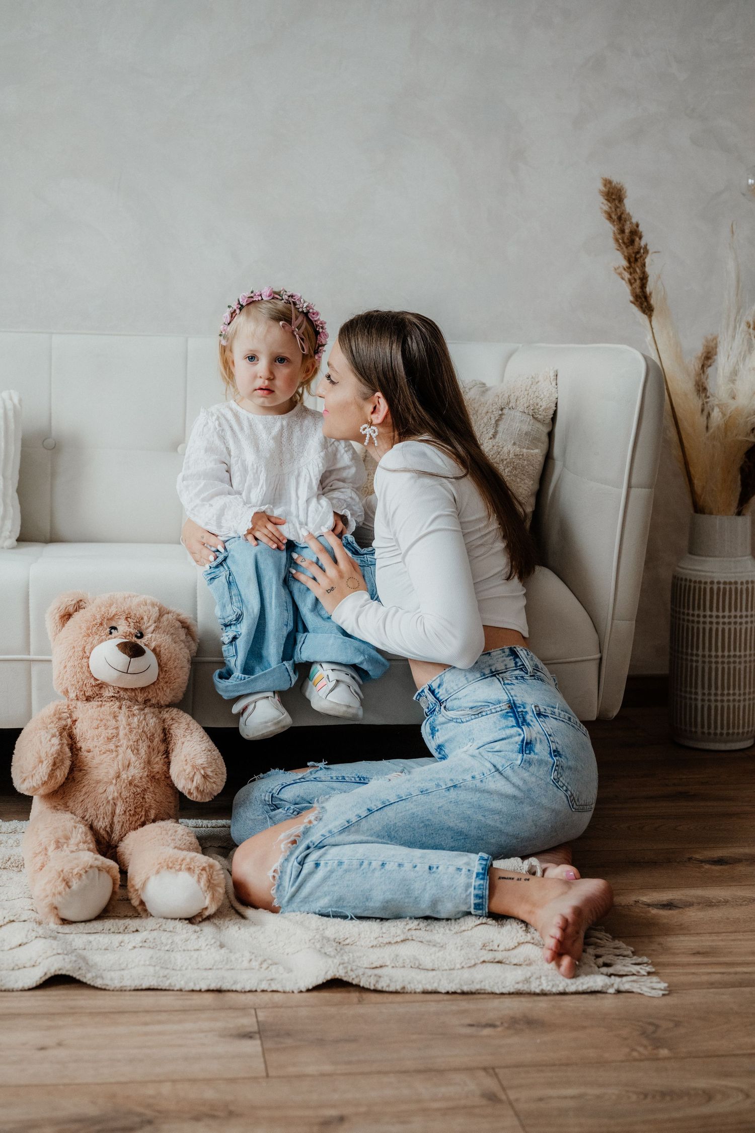 A mother in jeans kneels before her young daughter seated on a sofa, with a teddy bear on the floor nearby.