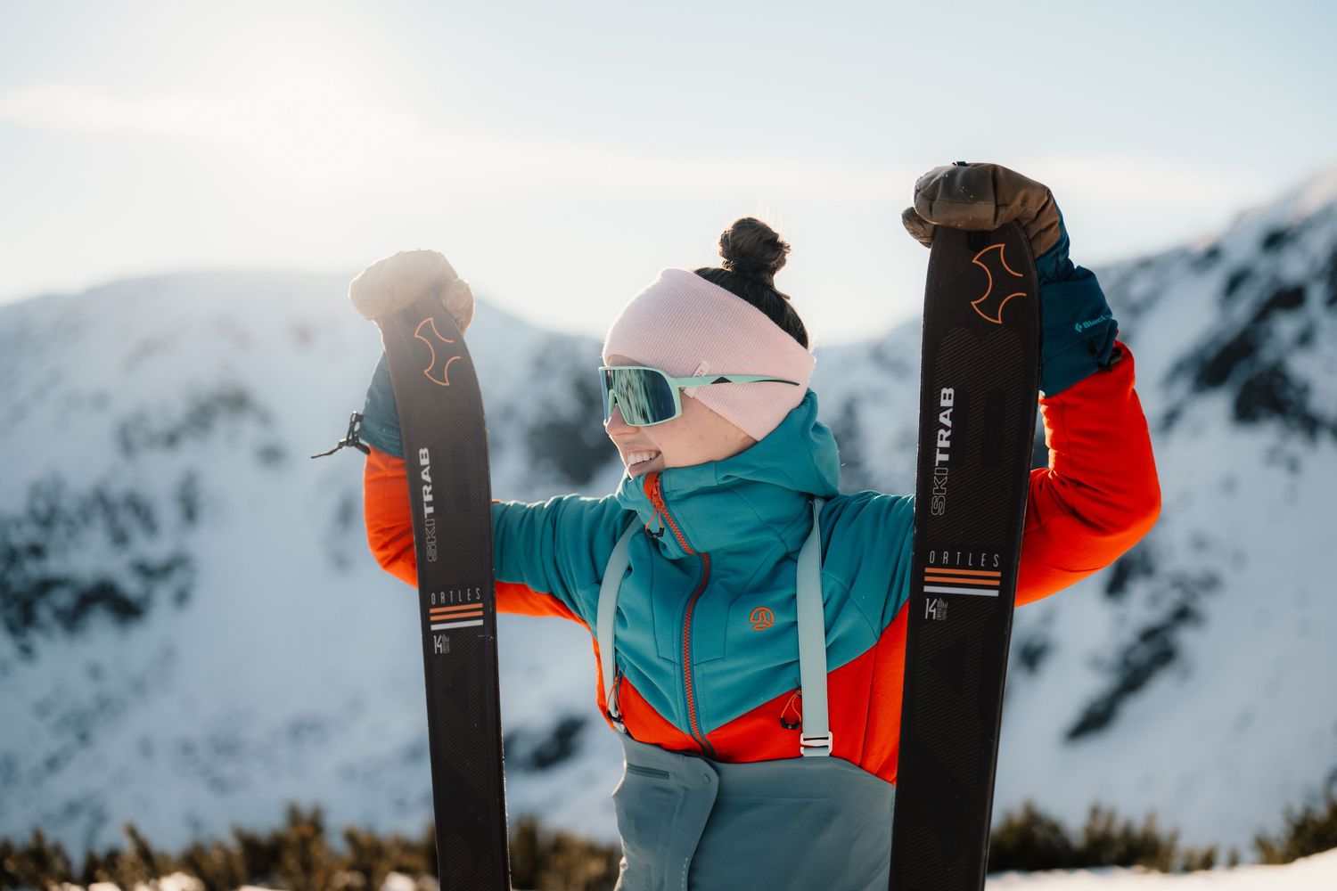Woman in teal and red ski jacket holding up skis against bright snowy mountain scenery.