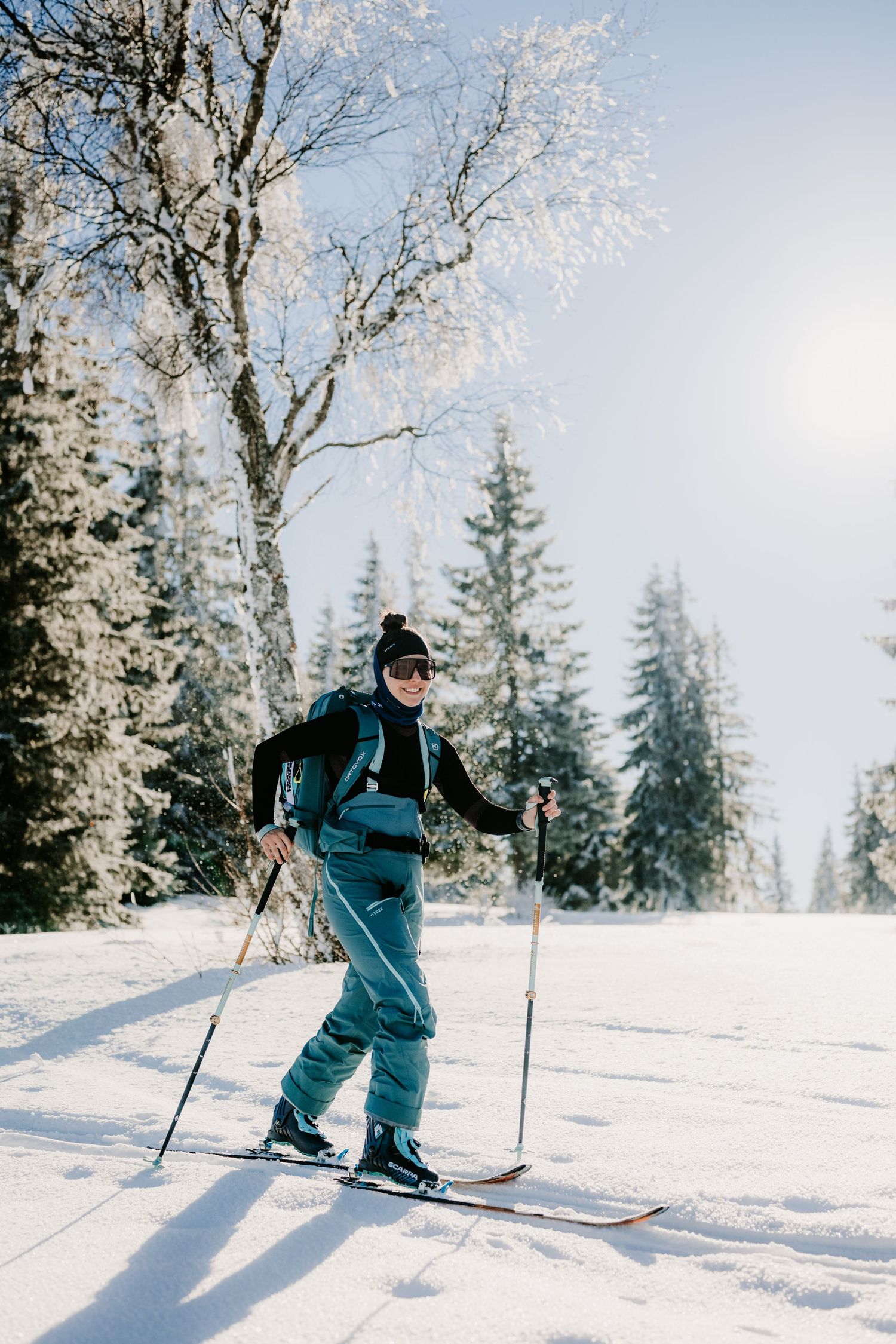 Female skier in teal outfit ski touring through a winter forest with frost-covered trees on sunny day.