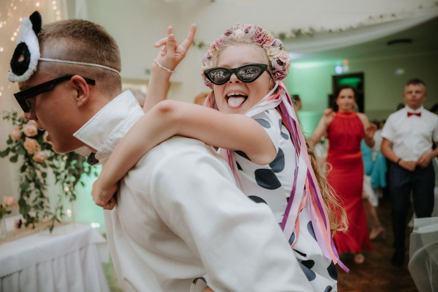 Bride and groom dancing joyfully at wedding reception wearing fun sunglasses, with guests celebrating in background.