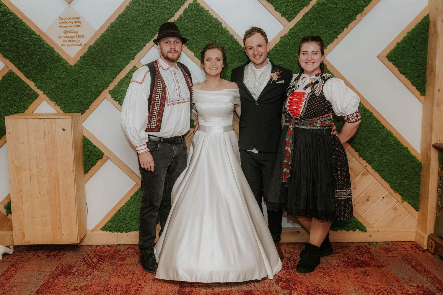 Wedding couple poses with two guests in traditional folk costumes before a decorative green moss wall backdrop.