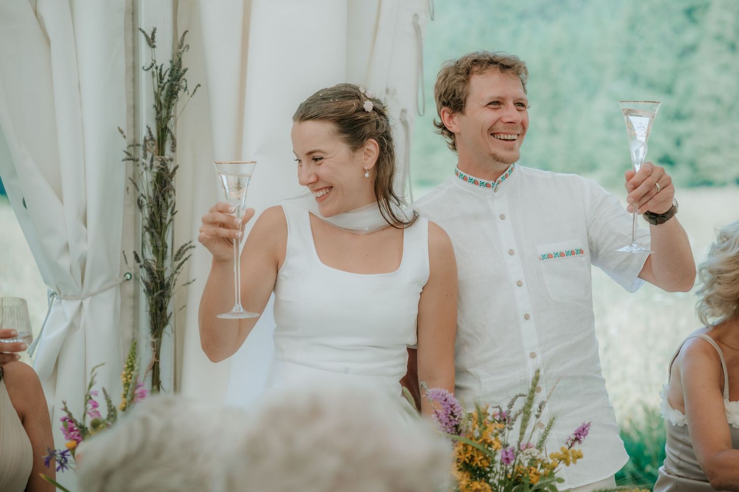 Happy newlyweds toasting with champagne glasses at outdoor wedding ceremony in white attire.