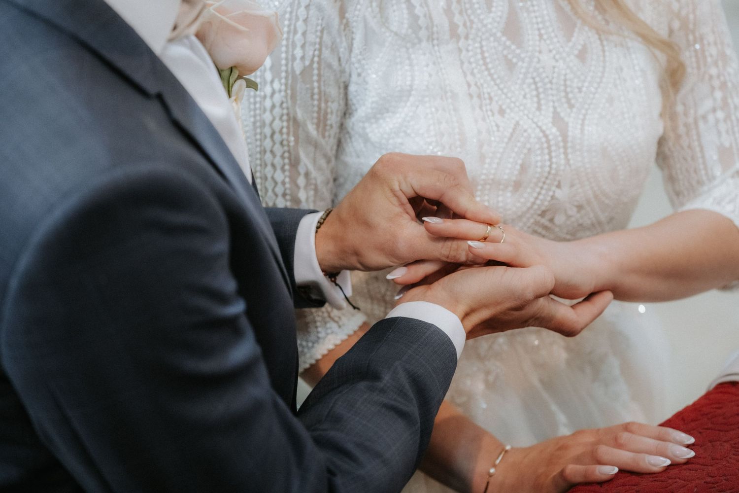 Bride placing a wedding ring on groom's finger during a ceremony, wearing a white lace dress.