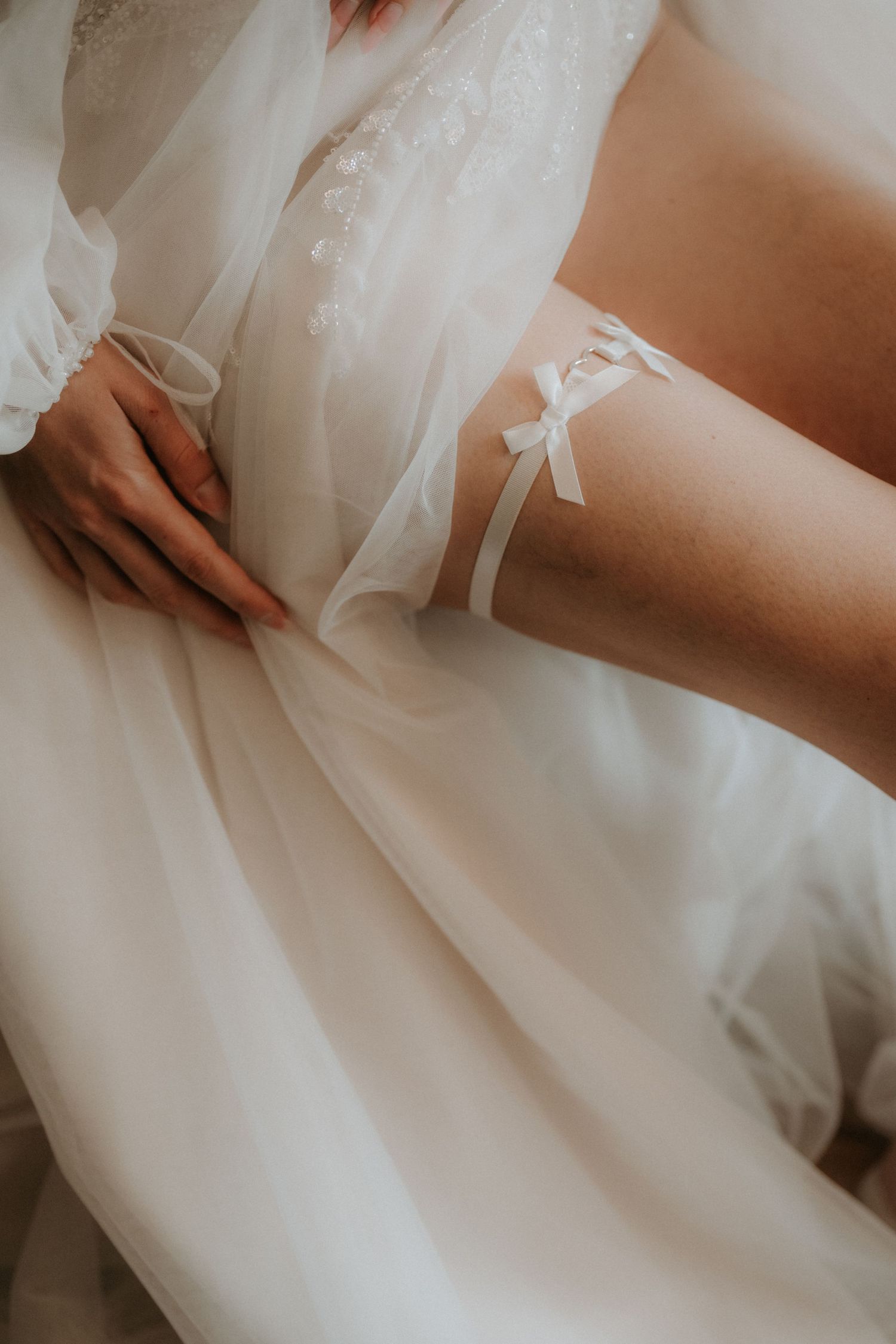 Bride wearing a white lace robe and delicate satin garter with bow on her thigh on wedding day.