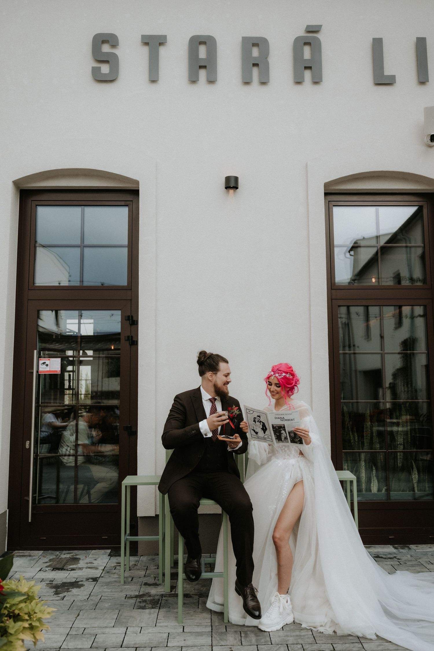 A bride with pink hair and groom in a suit sit outside Stara Li cafe, sharing a joyful moment on their wedding day.