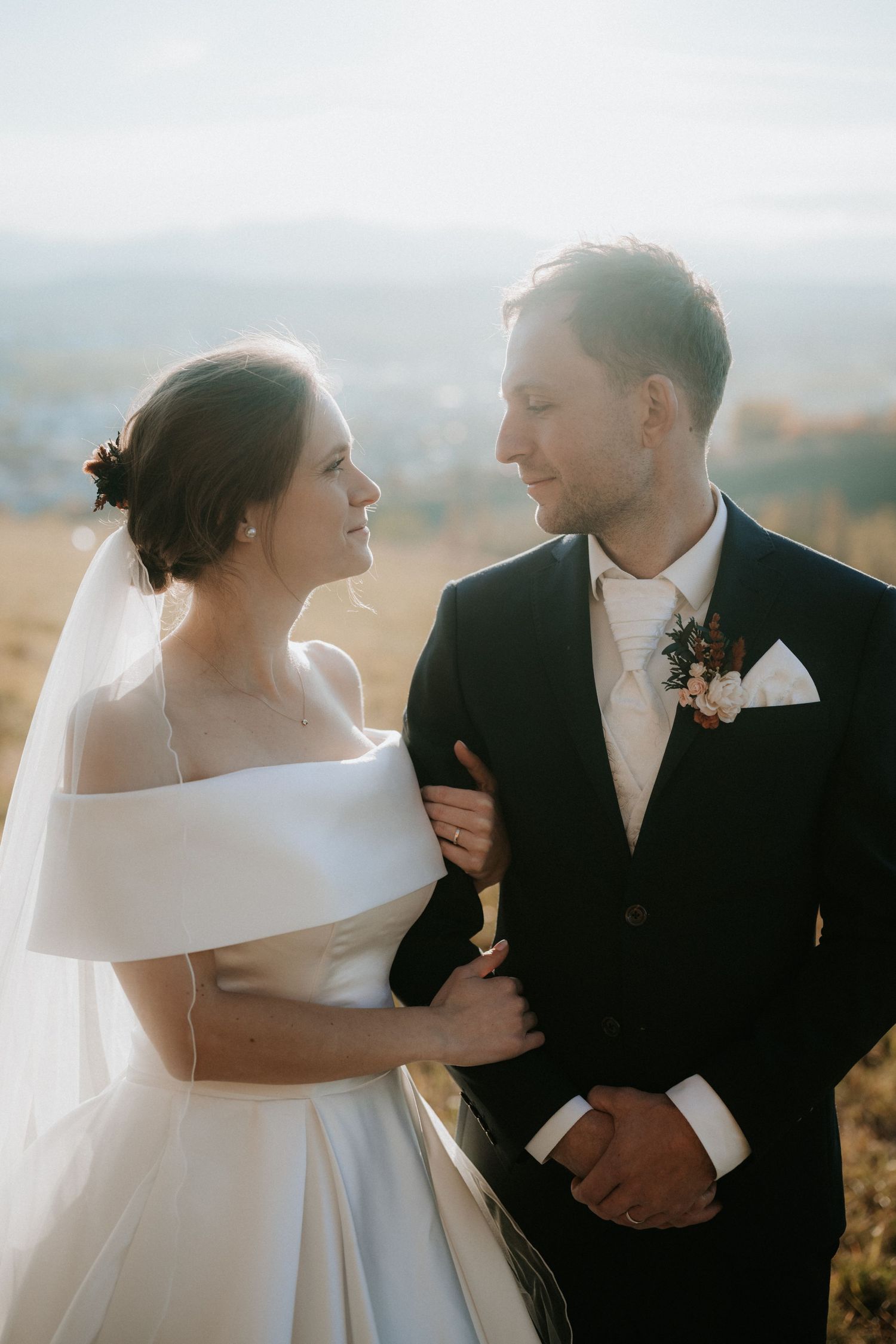 Bride and groom share a romantic moment outdoors, with the bride in an off-shoulder gown and groom in a dark suit with boutonniere.