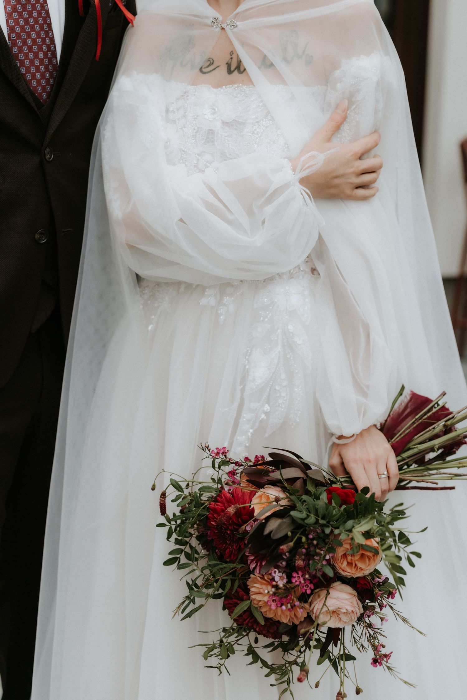 Bride in white lace gown with tulle cape holding a lush burgundy and blush wedding bouquet beside groom in dark suit.