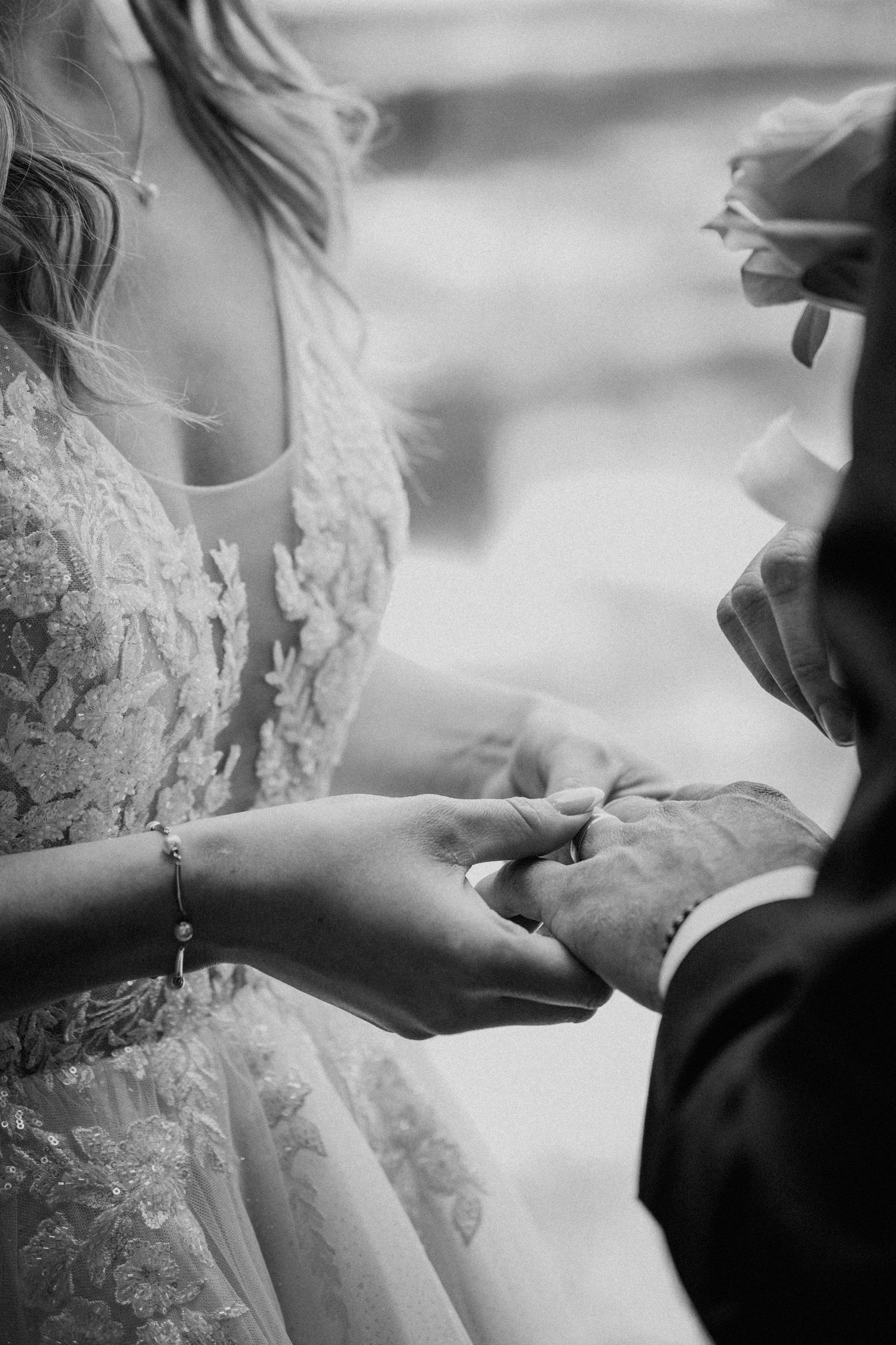Black and white photo of bride placing wedding ring on grooms finger during ceremony, wearing lace gown and bracelet.