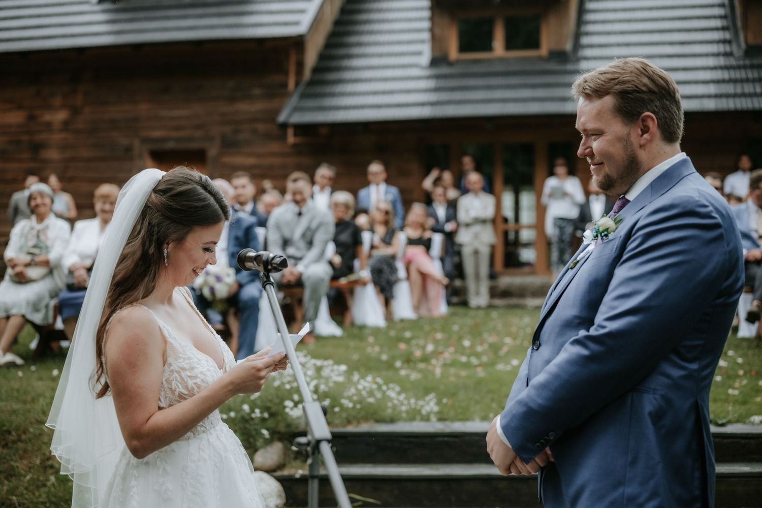 Bride reading vows to groom during outdoor wedding ceremony with guests seated in background near rustic wooden building.