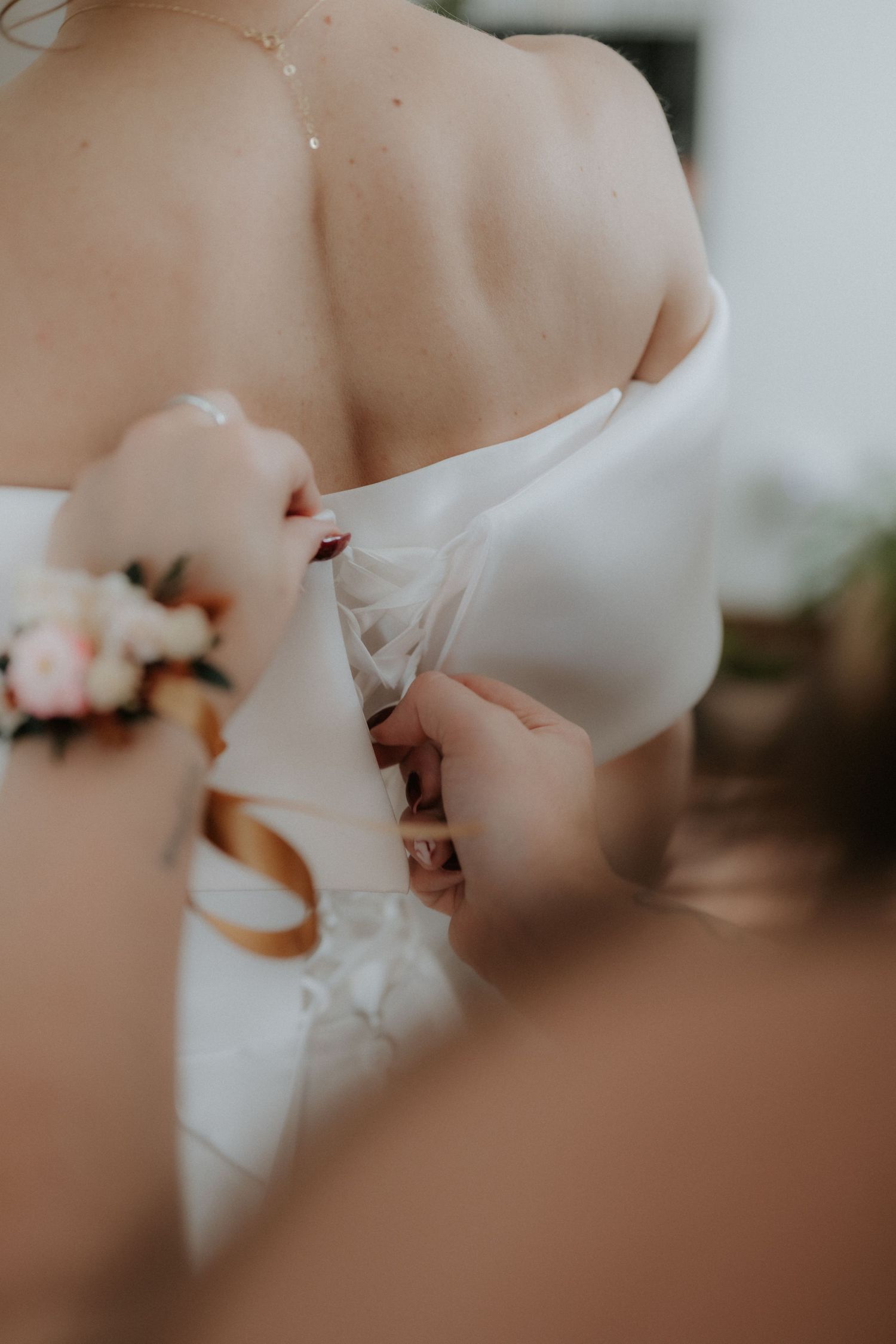 Bridesmaid fastening the back of a white off-shoulder wedding dress, with floral wrist corsage visible.