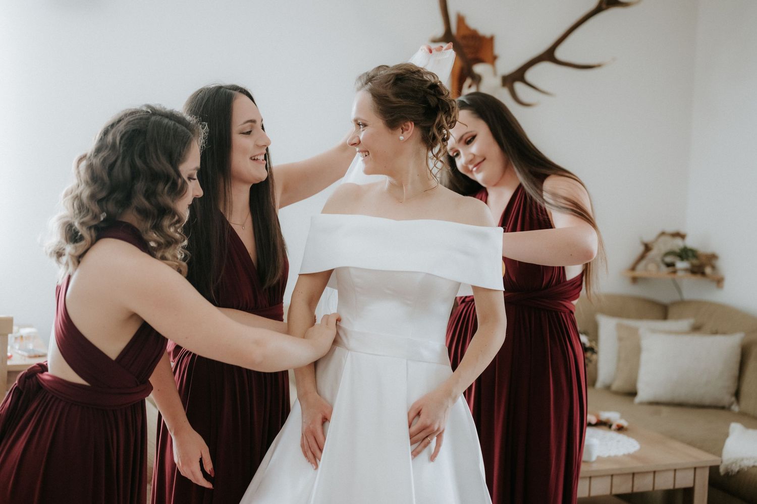Bridesmaids in burgundy dresses help bride in white off-shoulder gown prepare for wedding ceremony indoors.