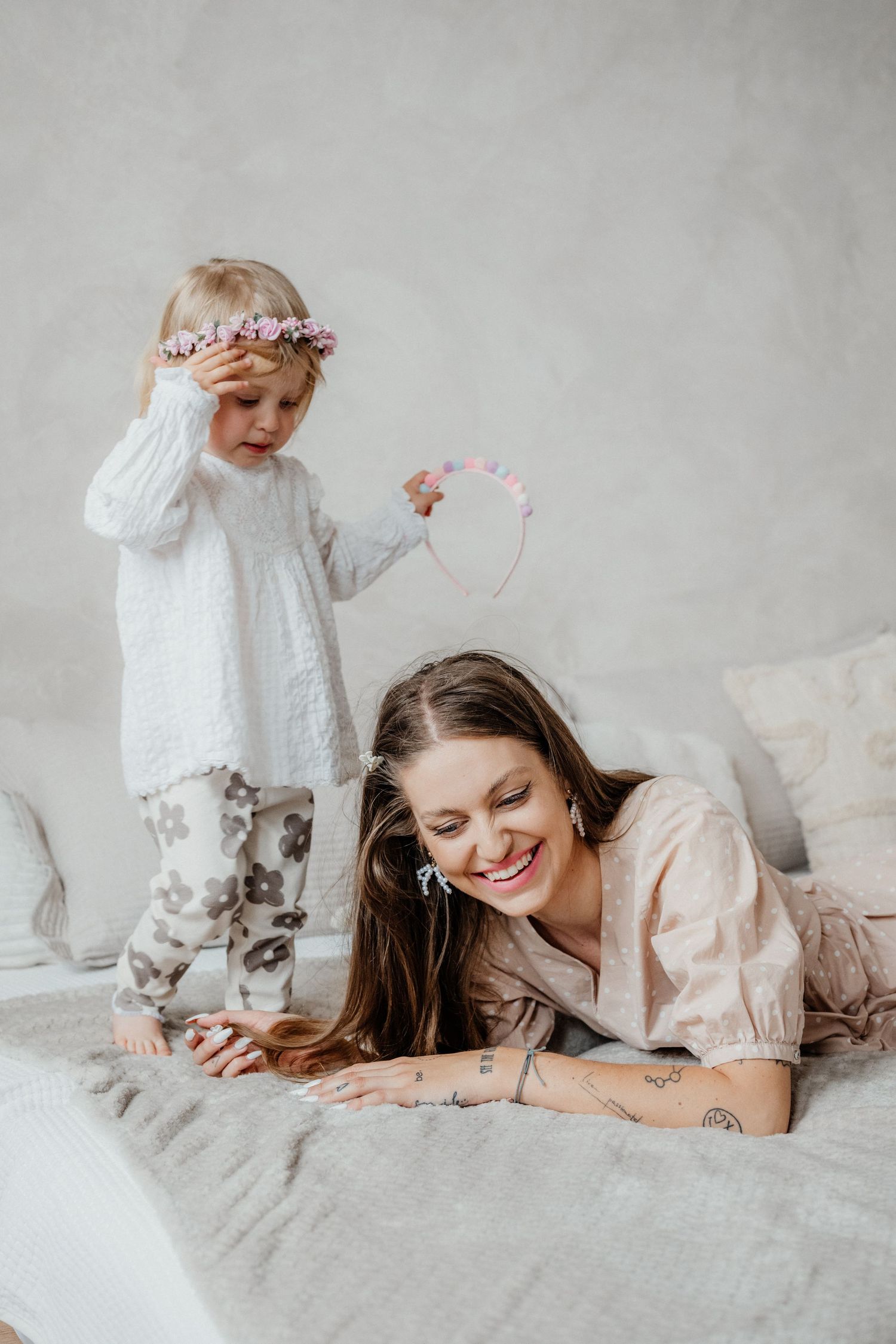 A smiling mother lying on a bed as her toddler daughter places a flower crown on her head.