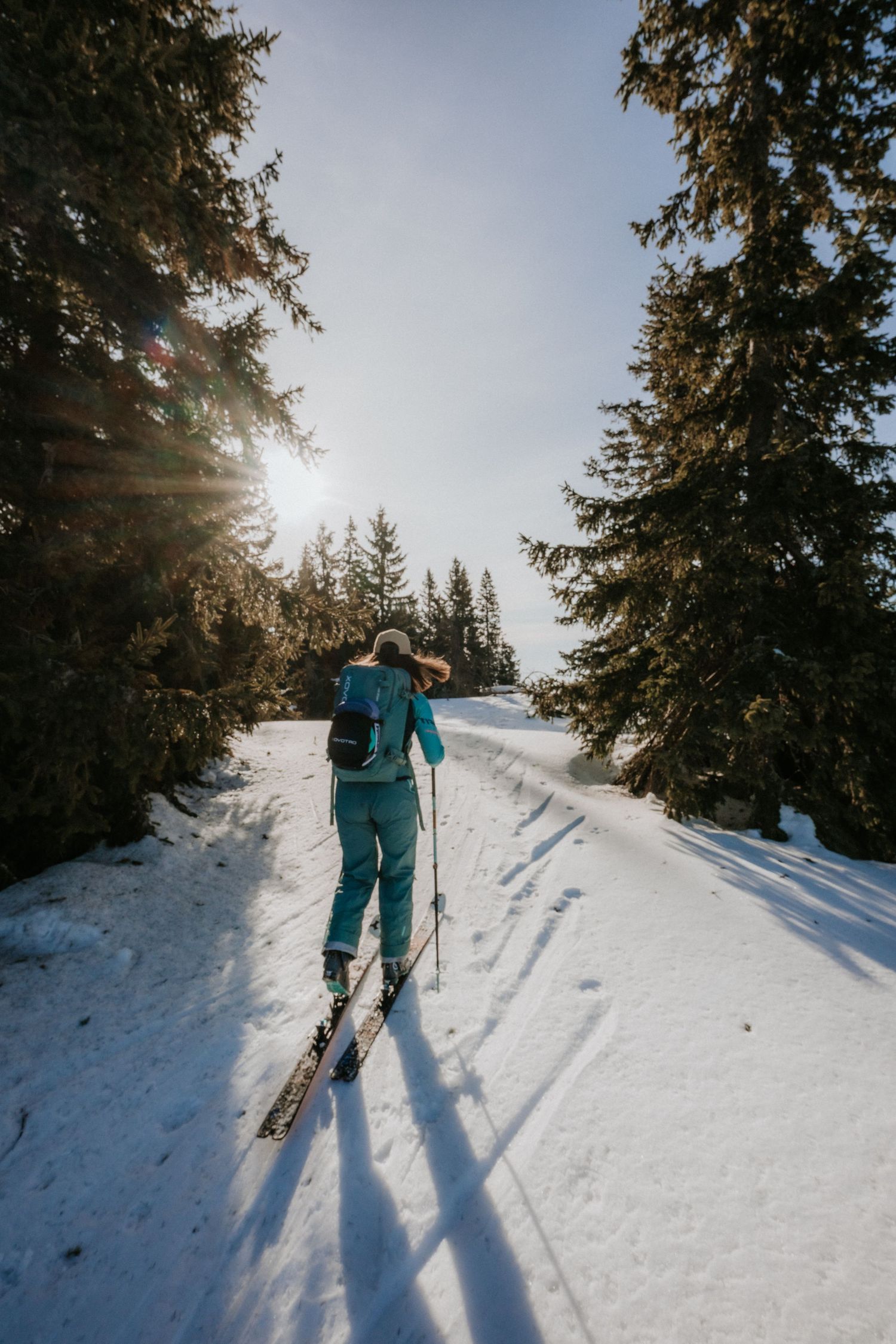 Female skier in teal suit ascending snowy forest trail with sunlight filtering through pine trees.