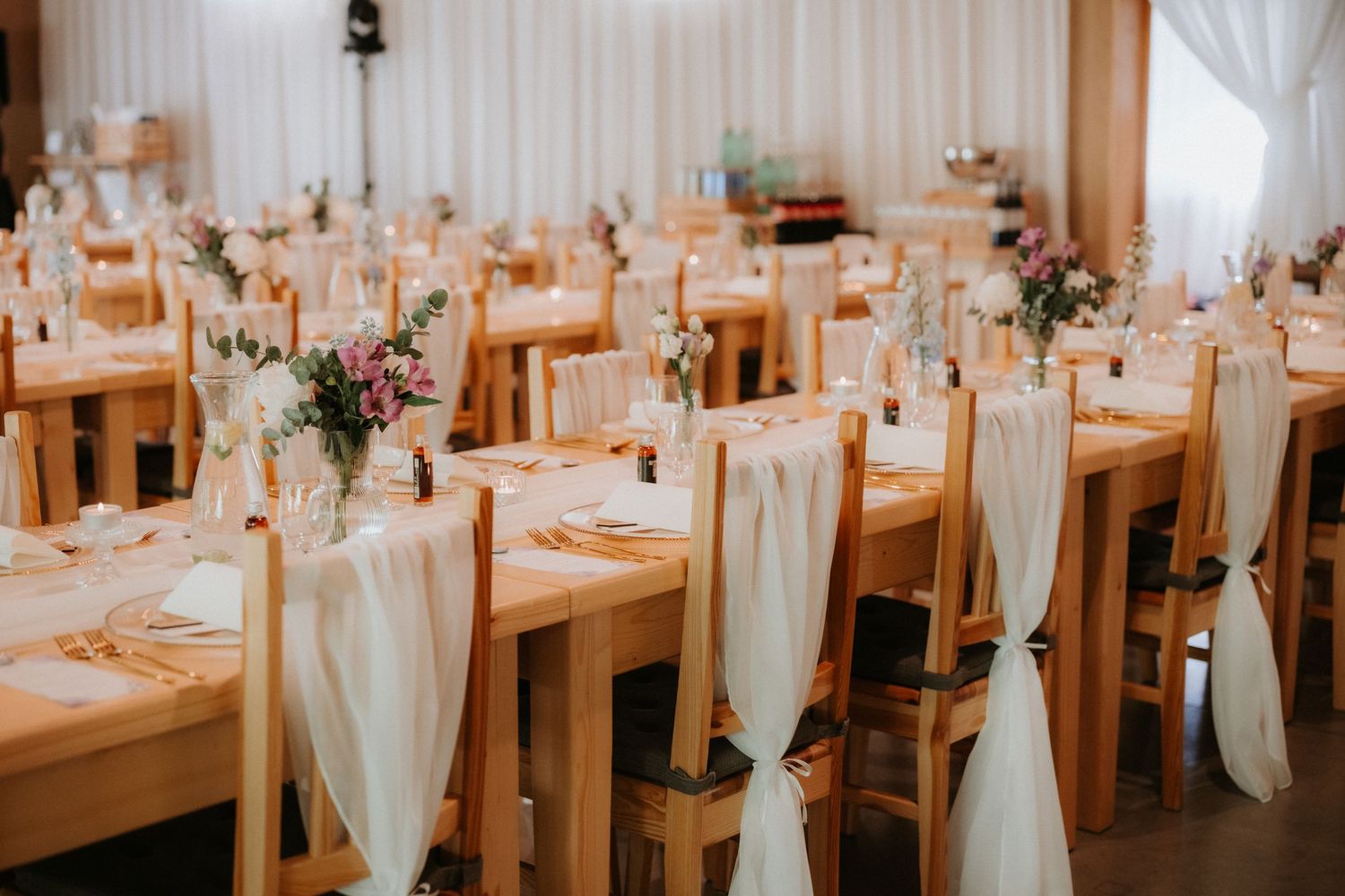 Elegant wedding reception hall with wooden chairs adorned with white sashes and floral centerpieces on decorated tables.
