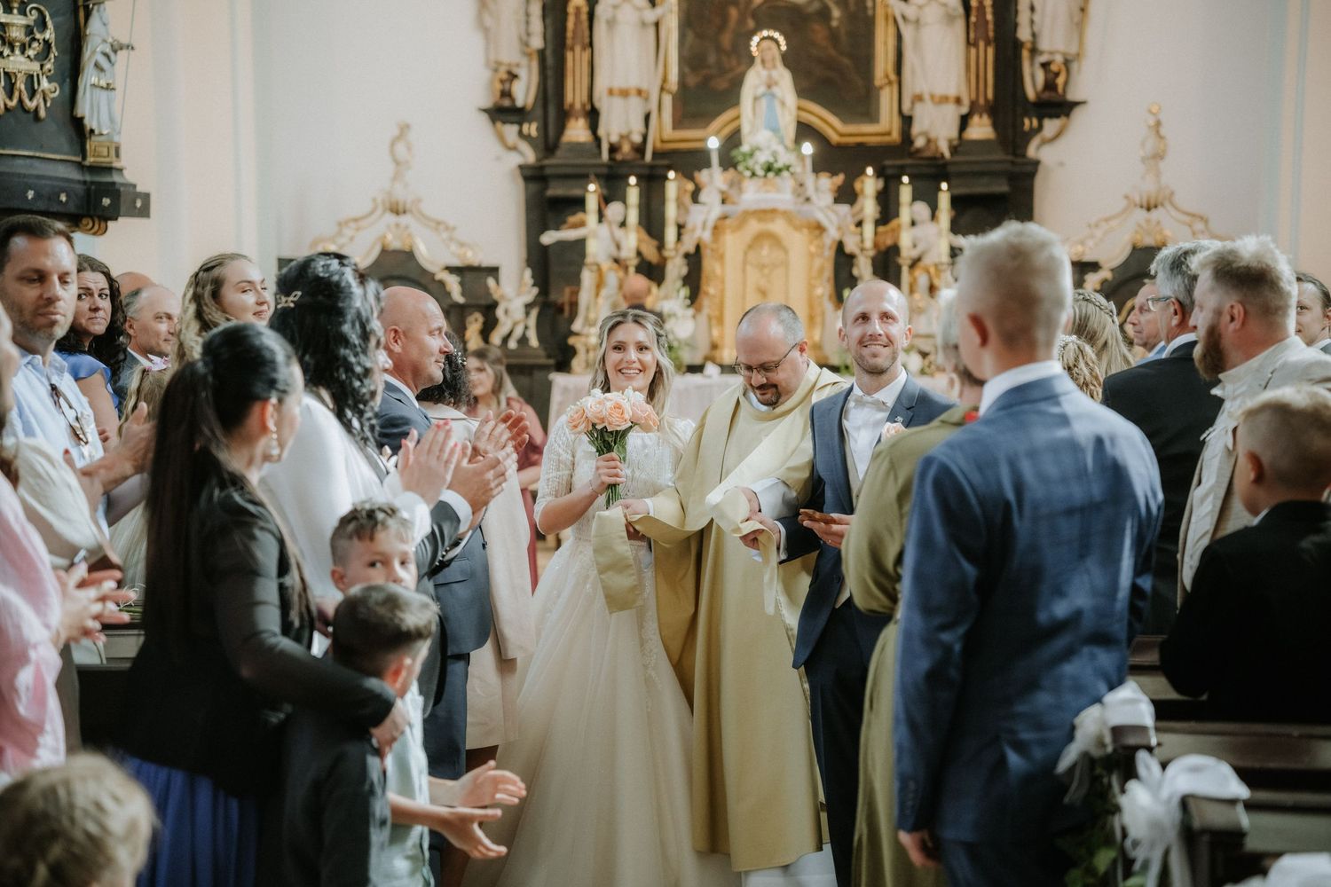 Bride and groom walk down church aisle surrounded by applauding guests during a traditional wedding ceremony.