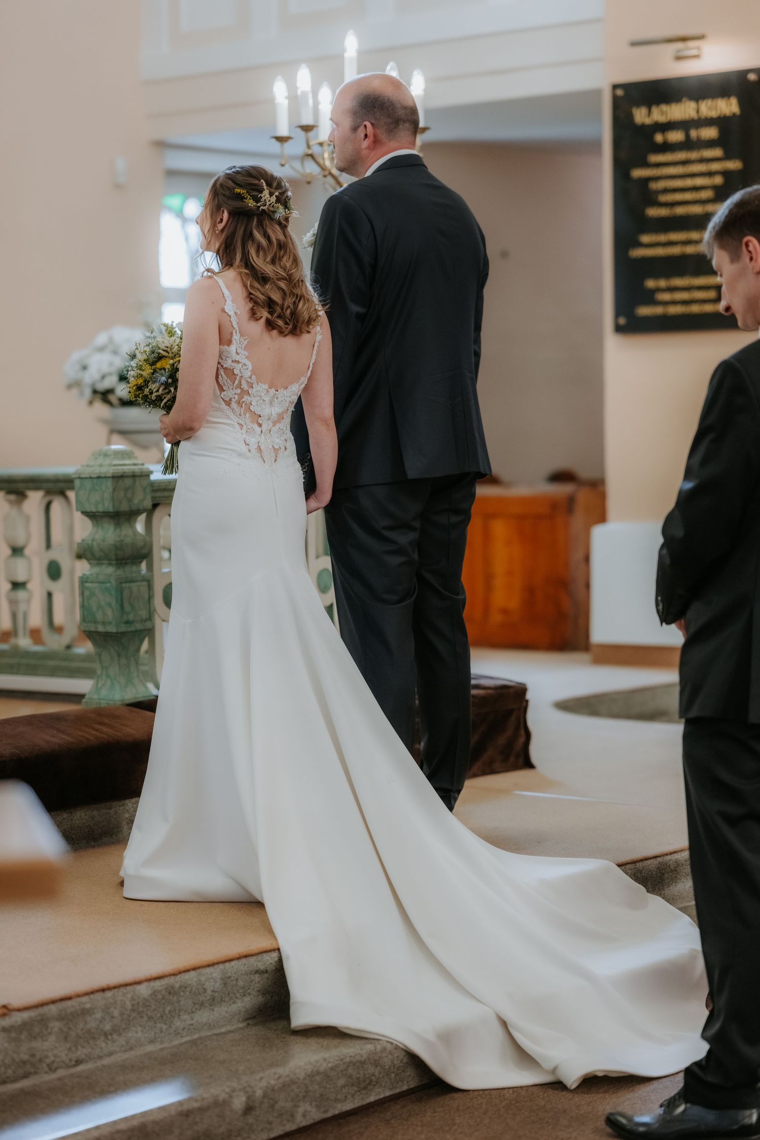 Bride in white lace gown and groom in black suit stand at church altar during wedding ceremony.