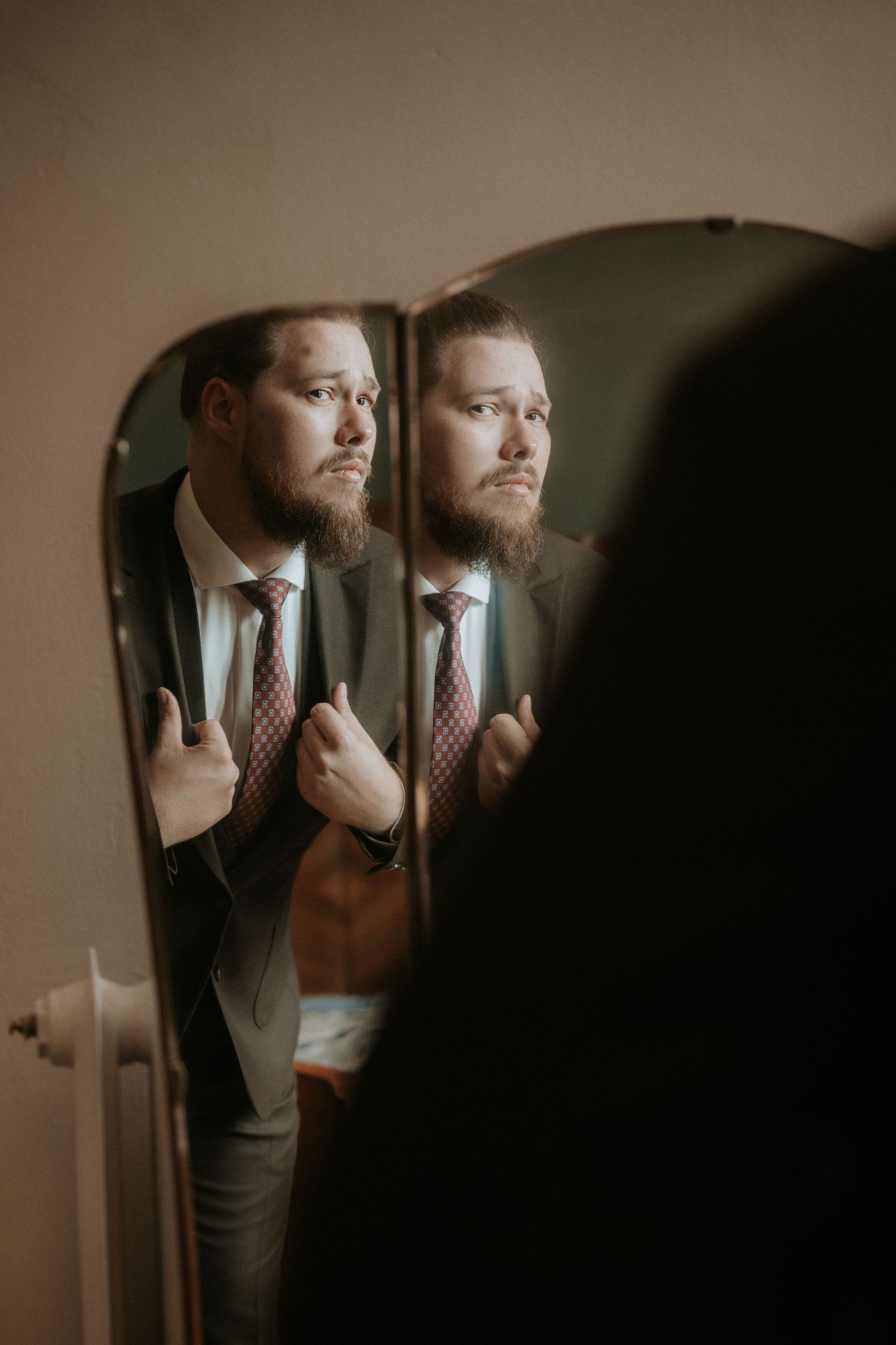 Bearded groom in gray suit adjusting tie while looking at his reflection in a vintage mirror.