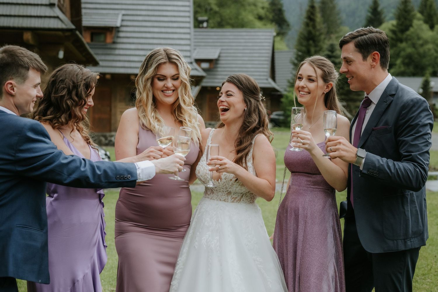 Bride in white gown laughs with bridesmaids in purple dresses and groomsmen holding champagne flutes outdoors.