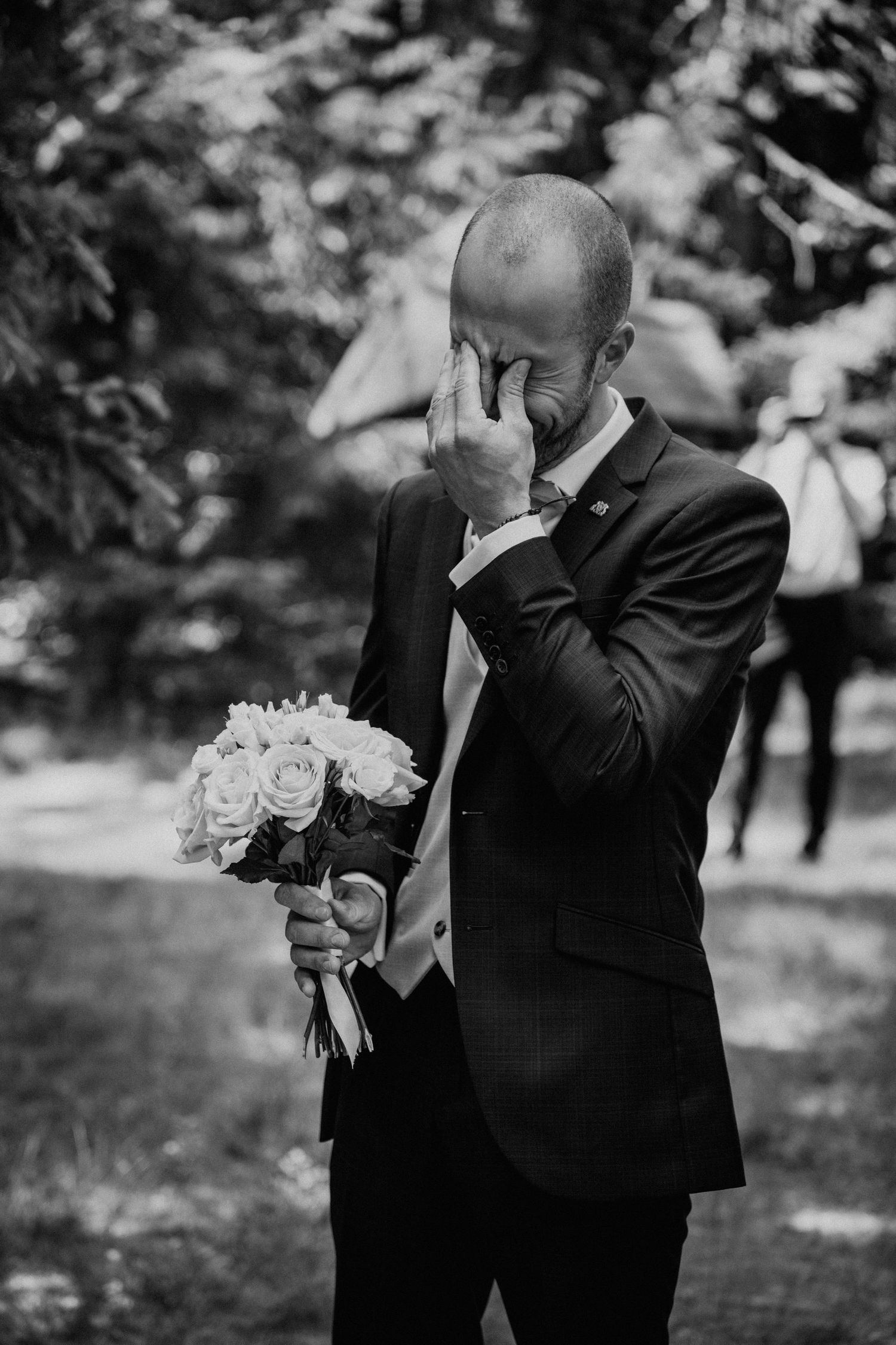 Emotional groom in black suit crying while holding bridal bouquet of roses outdoors in black and white photo.