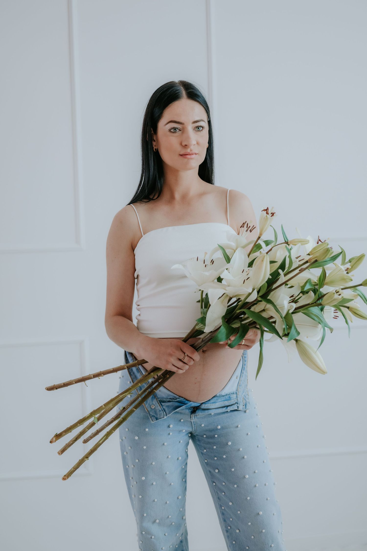 Pregnant woman in white crop top and pearl-embellished jeans holding white lily bouquet.