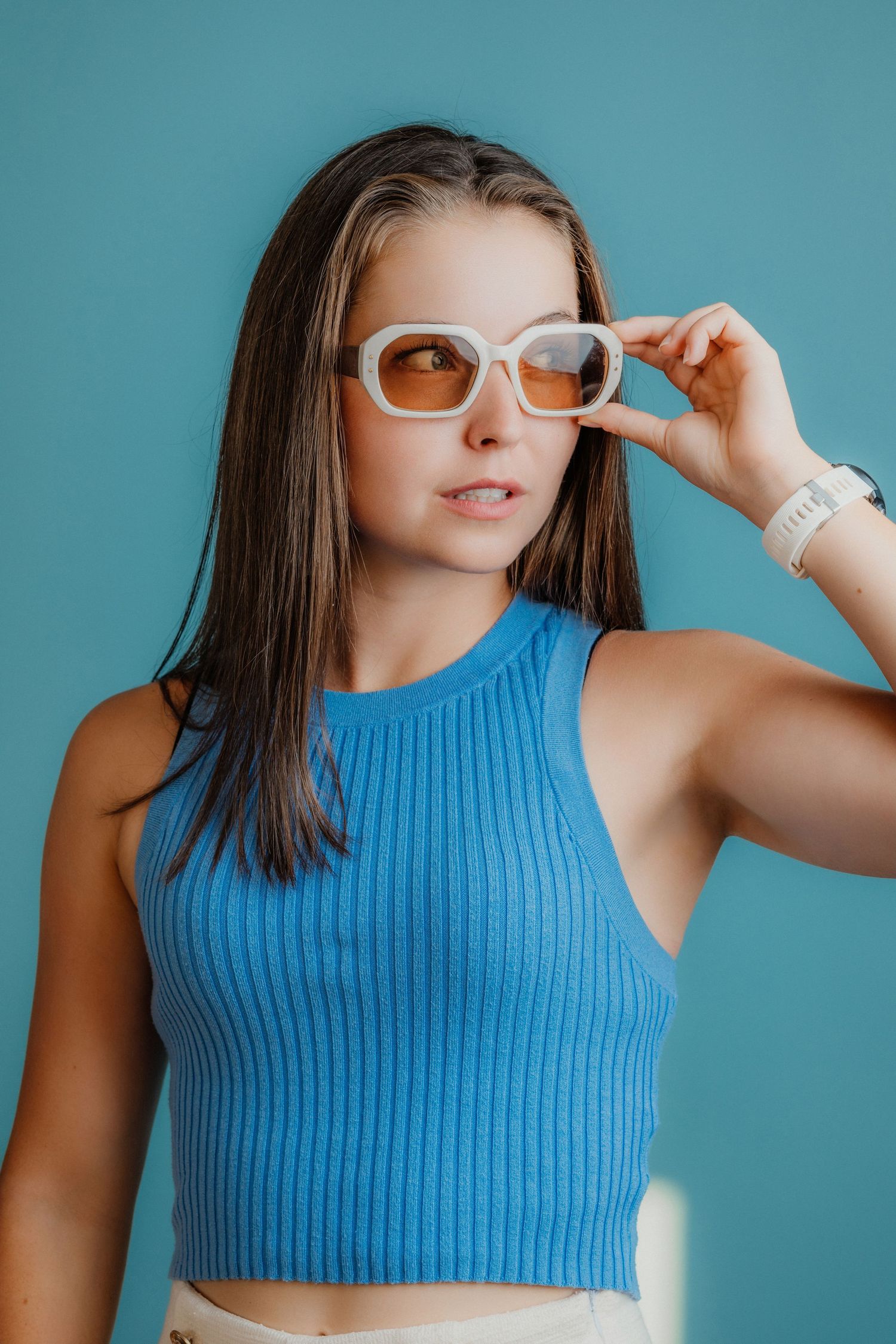 Young woman in blue ribbed crop top adjusting white sunglasses against teal background.